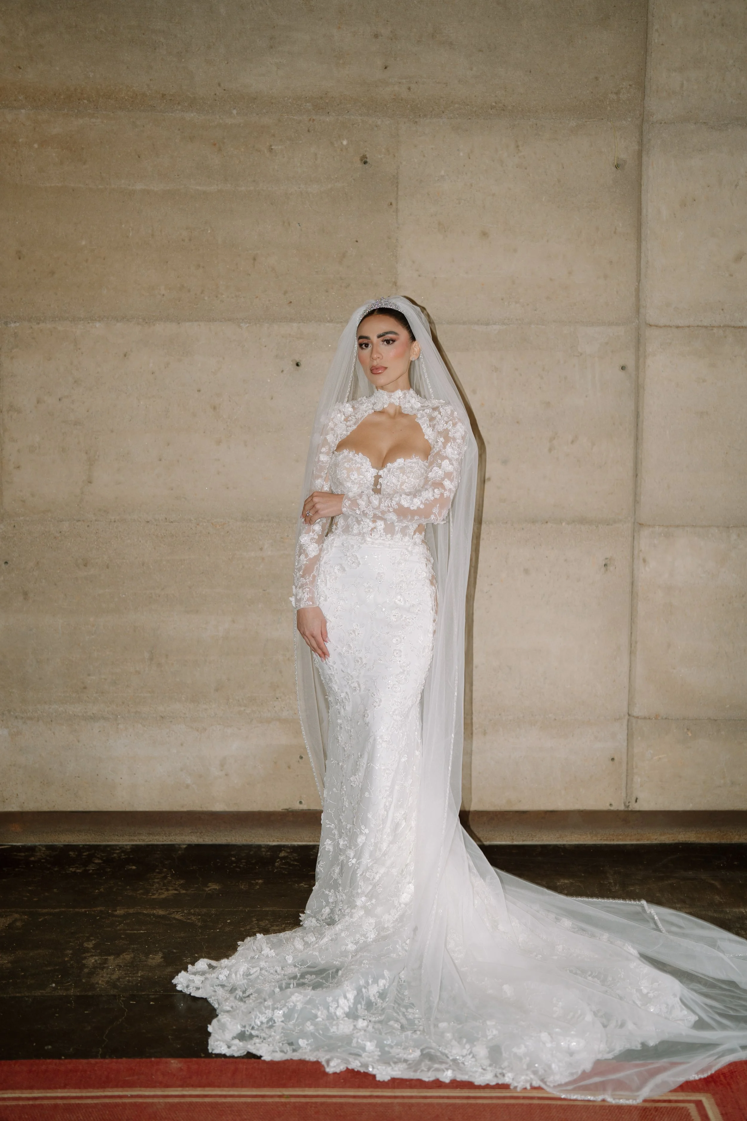 A bride in a white lace wedding gown with long sleeves, neckline, and veil, standing against a plain wall with her arms crossed. Wedding Photography in Valle de Guadalupe, Ensenada, Baja California.