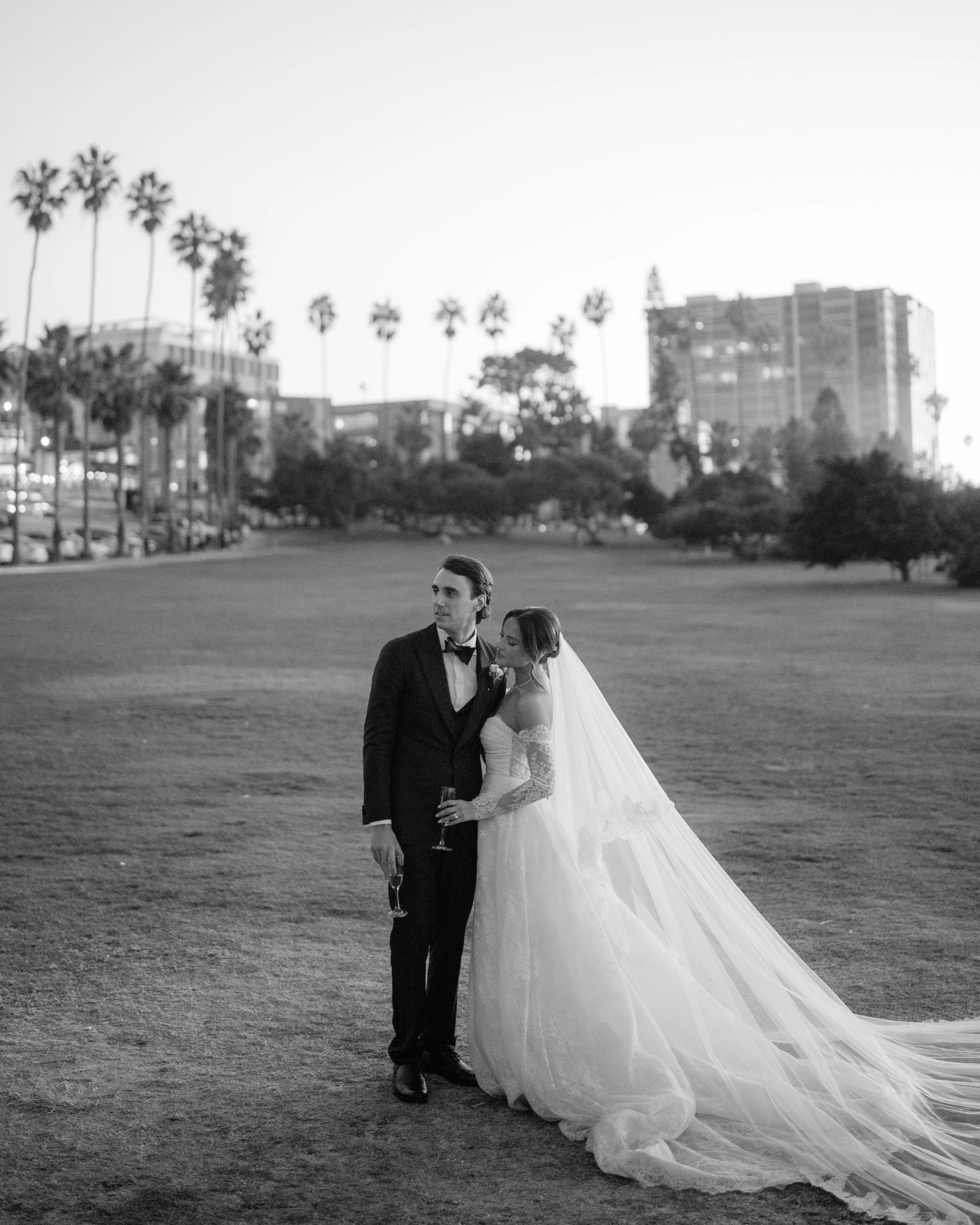 Black and white photo of a bride and groom standing on a grassy field, with the groom holding a glass of champagne and the bride leaning onto him, in an outdoor urban park setting. Wedding Photography in Valle de Guadalupe, Ensenada, Baja California.