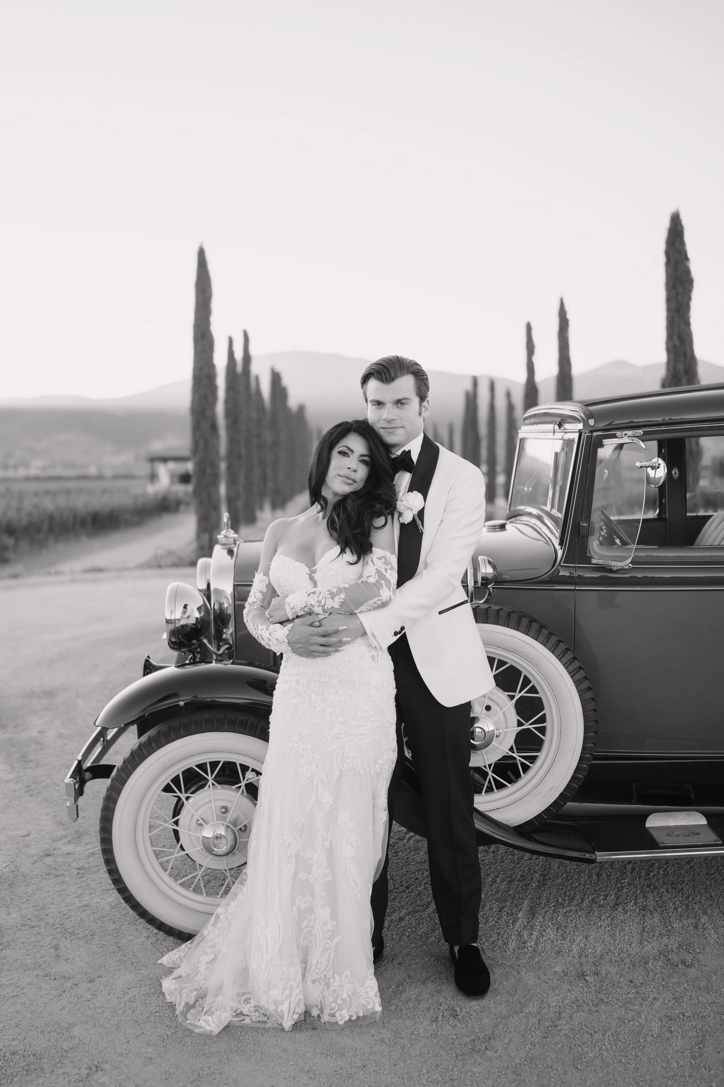 Black and white photo of a bride and groom standing together in front of a vintage car, with tall cypress trees and mountains in the background. Wedding Photography in Valle de Guadalupe, Ensenada, Baja California.