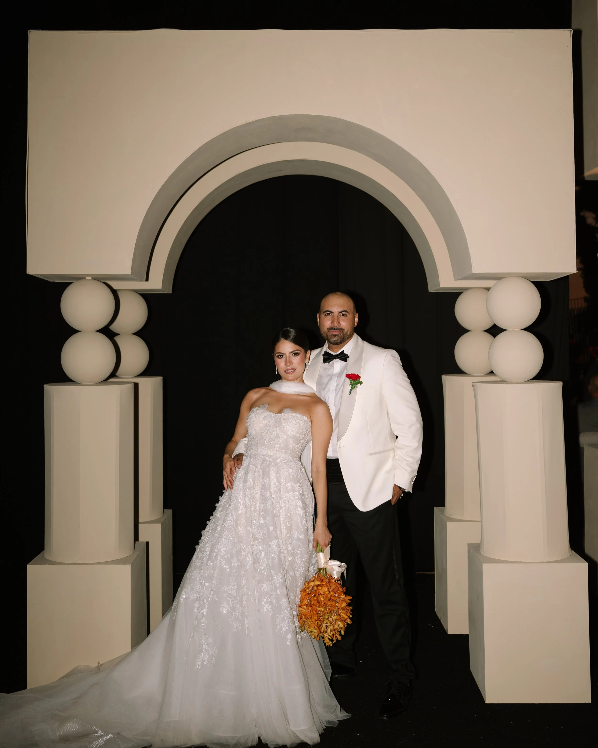 A bride and groom standing together in front of a decorative arch at their wedding. The bride is wearing a white wedding dress and the groom is in a tuxedo with a white jacket and black bow tie, holding a bouquet of orange flowers. Wedding Baja.