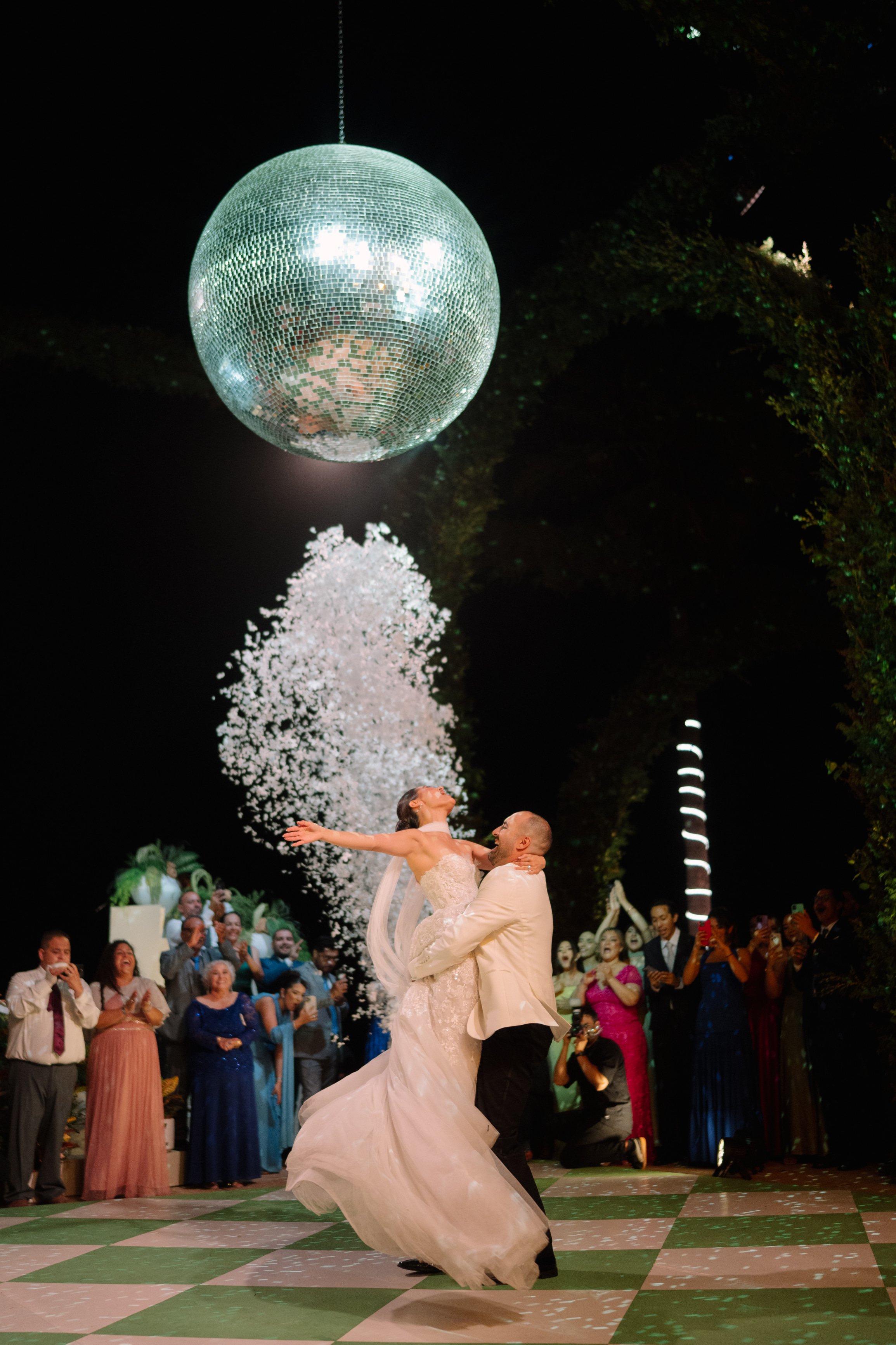 A bride and groom dancing at their wedding reception with a large disco ball overhead and guests watching and taking photos. Wedding Photography in Valle de Guadalupe, Ensenada, Baja California.
