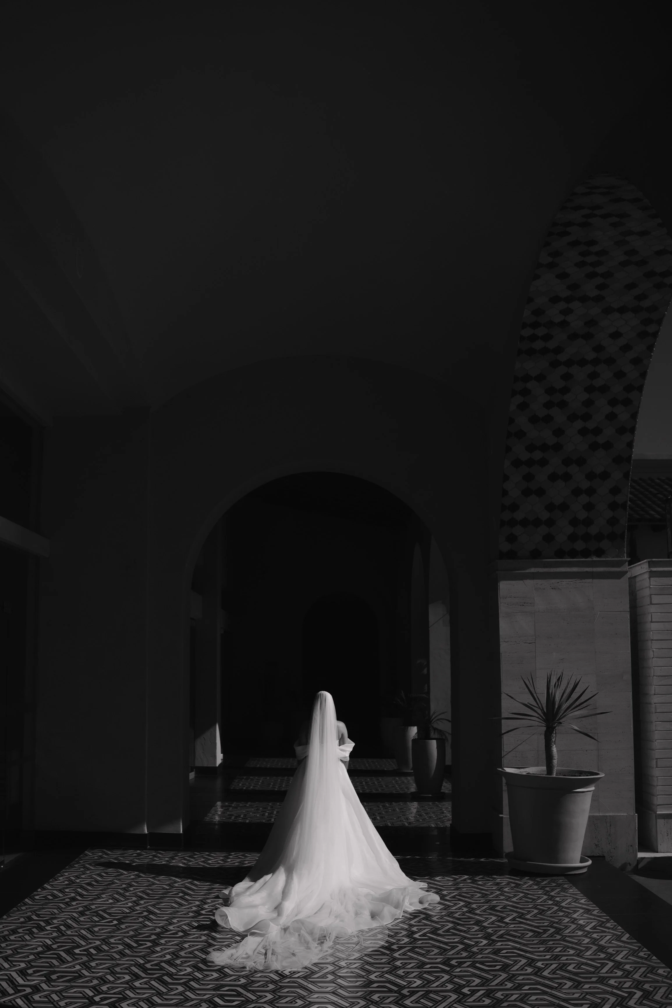A bride in a wedding dress standing with her back to the camera in a dimly lit indoor space with decorative planters and patterned tiles. Wedding Photography in Valle de Guadalupe, Ensenada, Baja California.