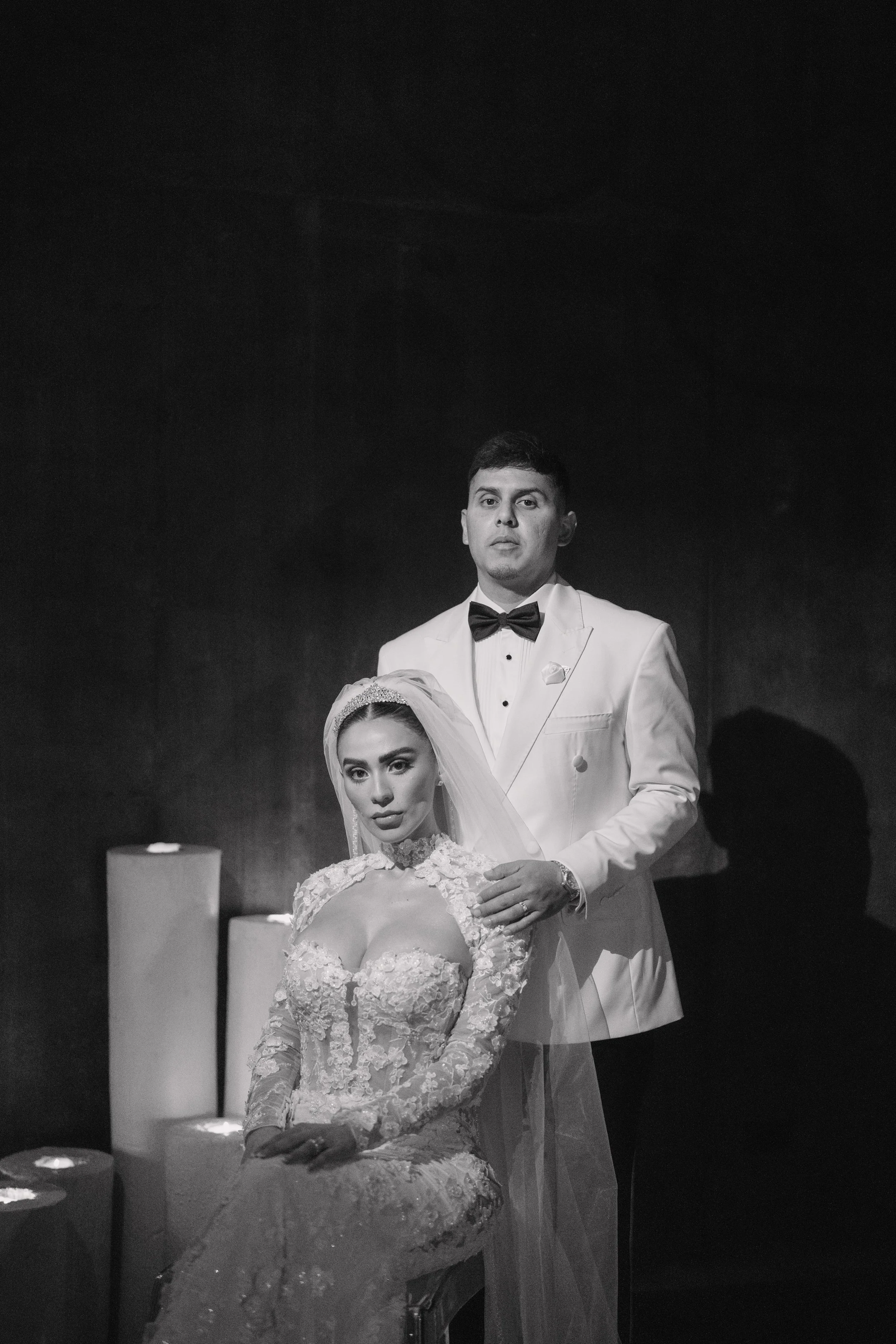 A black and white photo of a bride and groom in wedding attire. The bride is seated, wearing a lace wedding gown and veil, and the groom stands behind her in a white tuxedo with a black bow tie. Wedding Photography in Valle de Guadalupe, Ensenada.