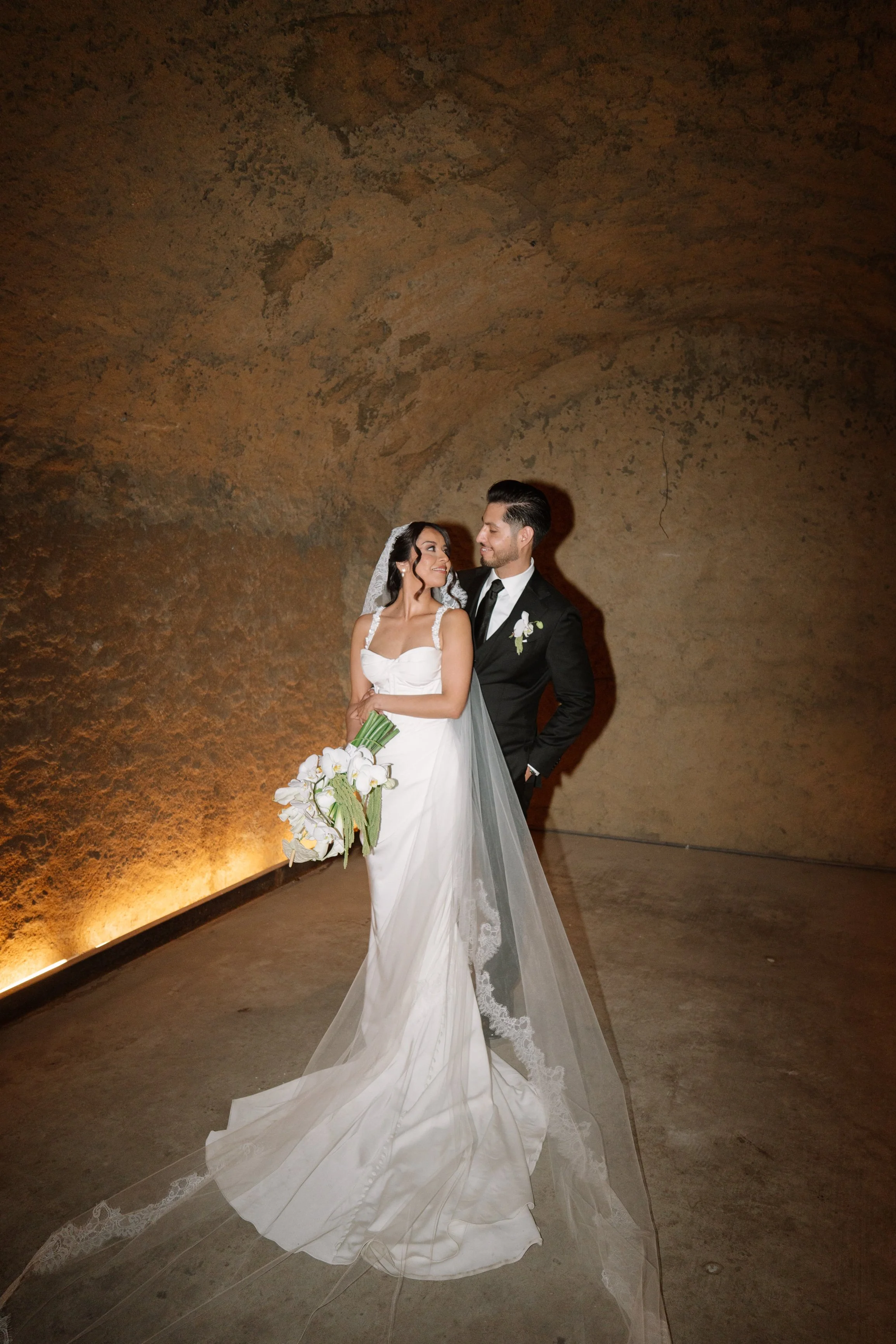 A bride in a white wedding gown holding a bouquet of white flowers, and a groom in a black tuxedo with a white boutonniere, standing in a dimly lit, rustic setting. Wedding Photography in Valle de Guadalupe, Ensenada, Baja California.