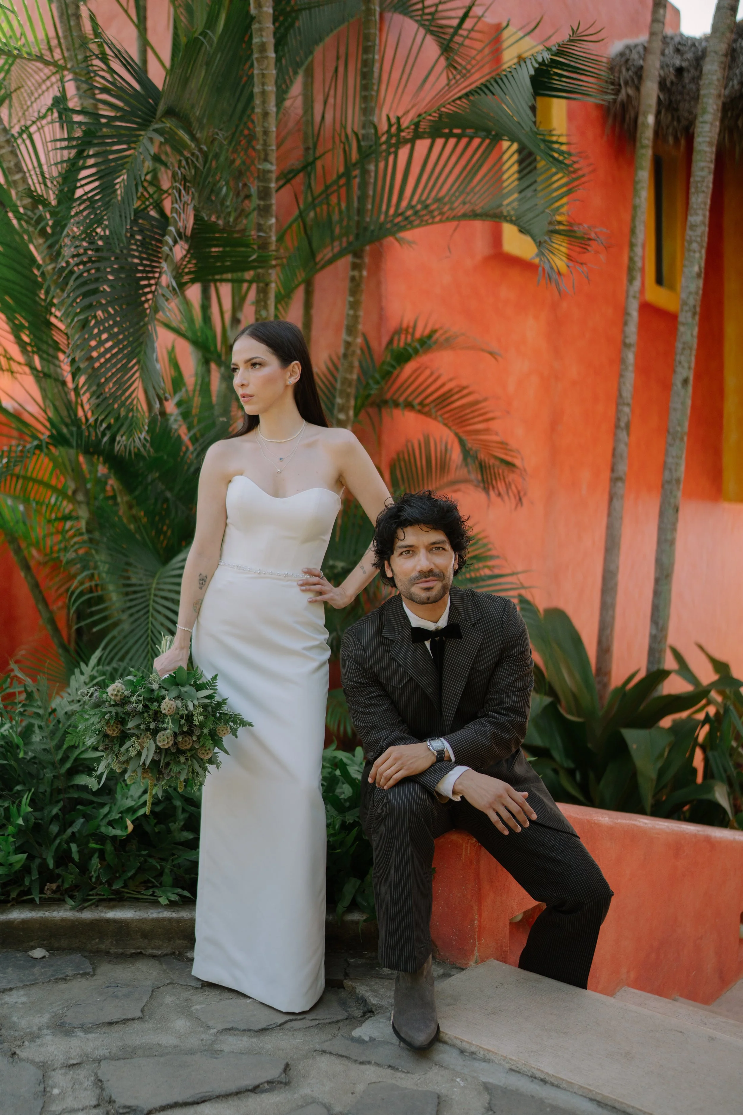 A woman in a white wedding dress holding a bouquet standing next to a man in a black suit and bow tie sitting on a low wall, with tropical plants and an orange wall in the background. Wedding Photography in Valle de Guadalupe, Ensenada, Baja Californ