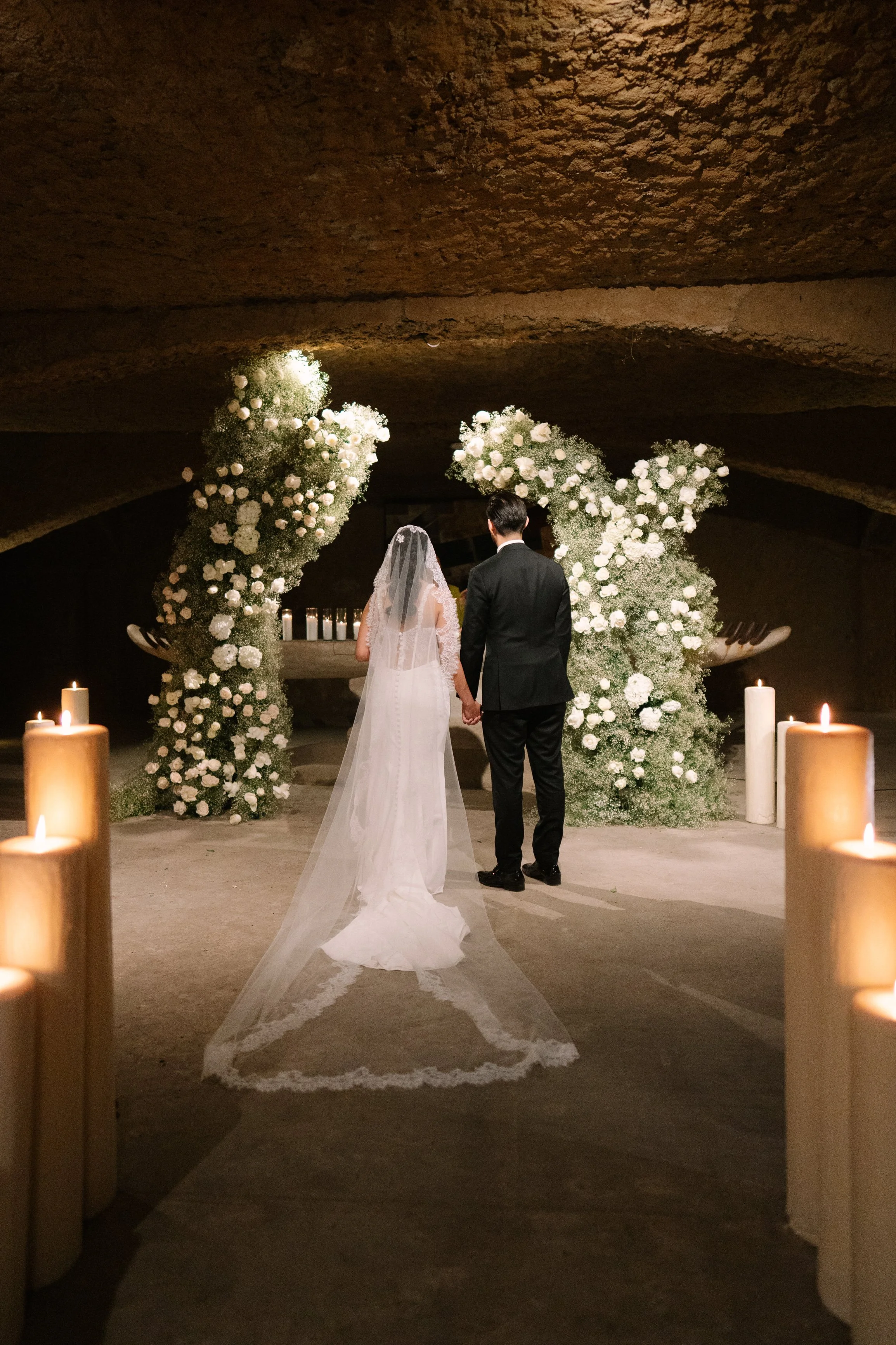 Bride and groom holding hands at wedding ceremony under floral arch with candles surrounding them. Wedding Photography in Valle de Guadalupe, Ensenada, Baja California.