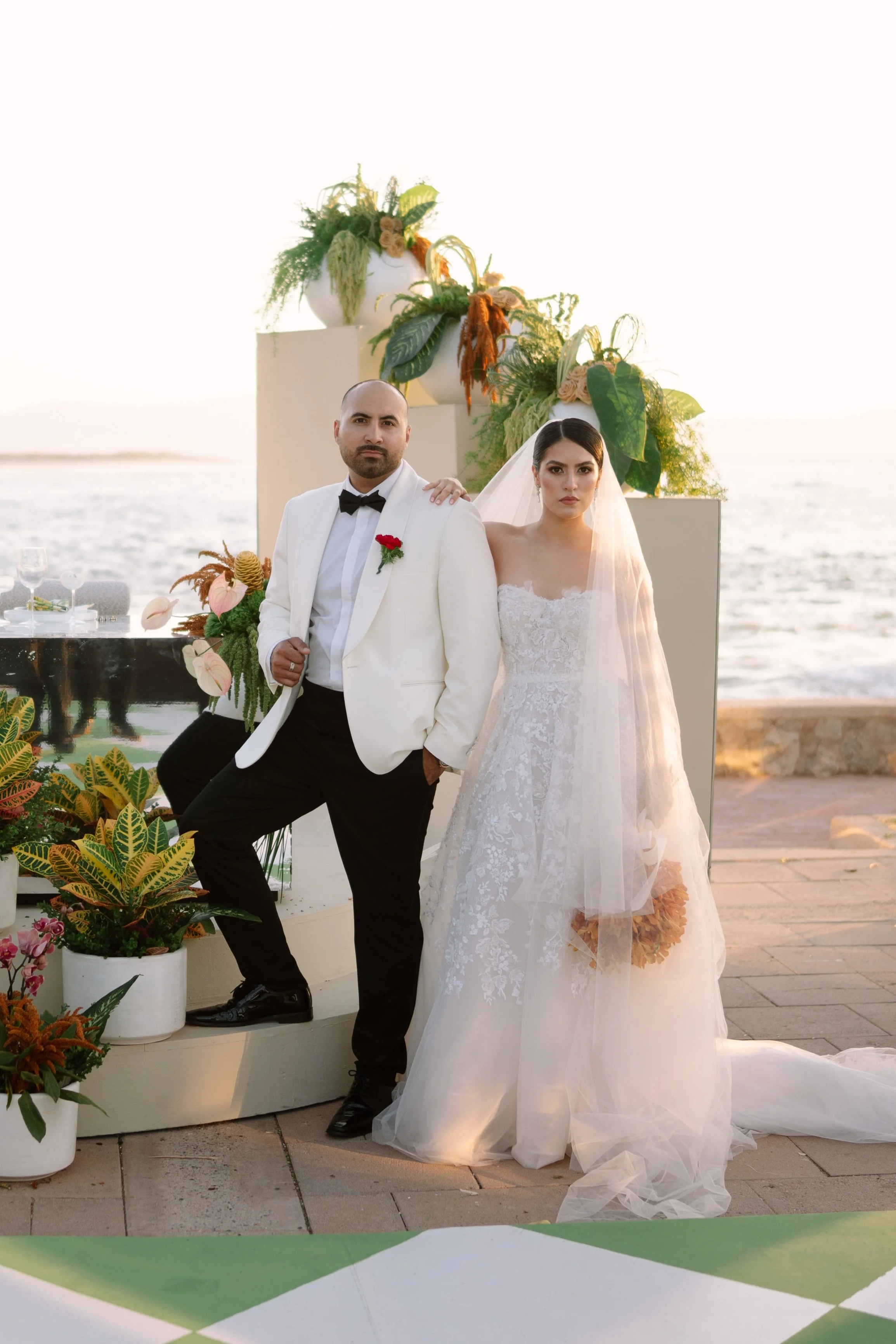 A bride and groom stand together on a seaside wedding altar with lush greenery and floral arrangements, during sunset. Wedding Photography in Valle de Guadalupe, Ensenada, Baja California.