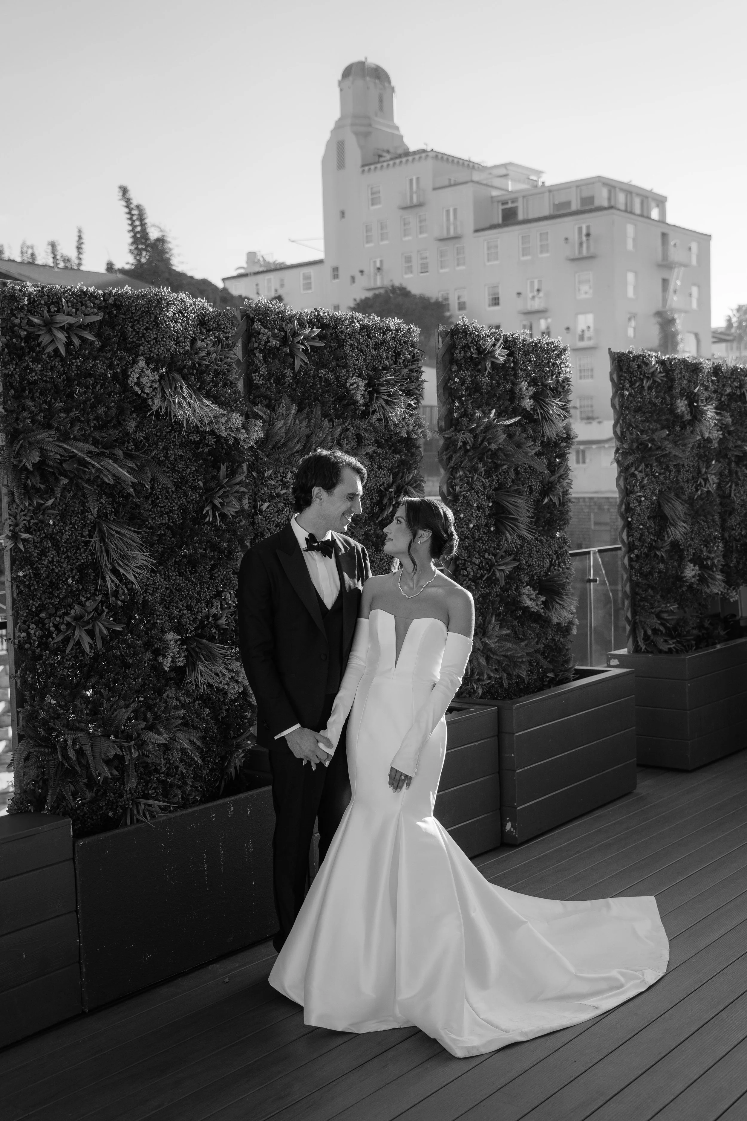 Black and white photo of a bride and groom holding hands, looking at each other, standing outdoors with a building and greenery behind them. Wedding Photography in Valle de Guadalupe, Ensenada, Baja California.