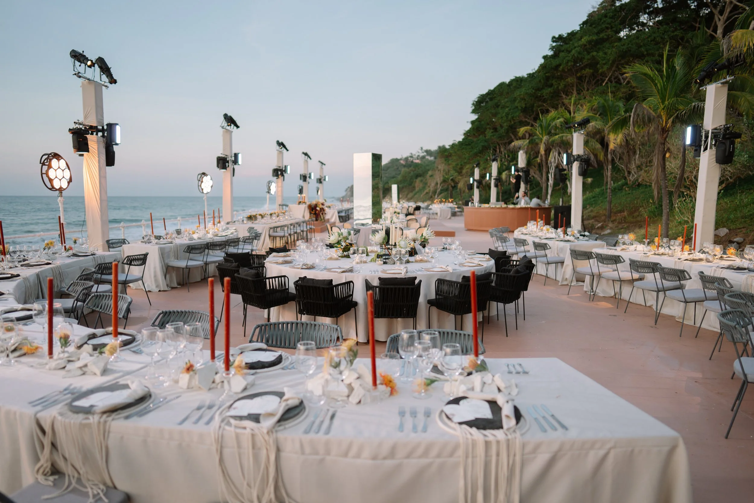 Set up for an outdoor beachside event with round and rectangular tables covered with white tablecloths, decorated with flowers, candles, and tableware, under lighting fixtures, overlooking the ocean with lush trees in the background at dusk. Baja. 