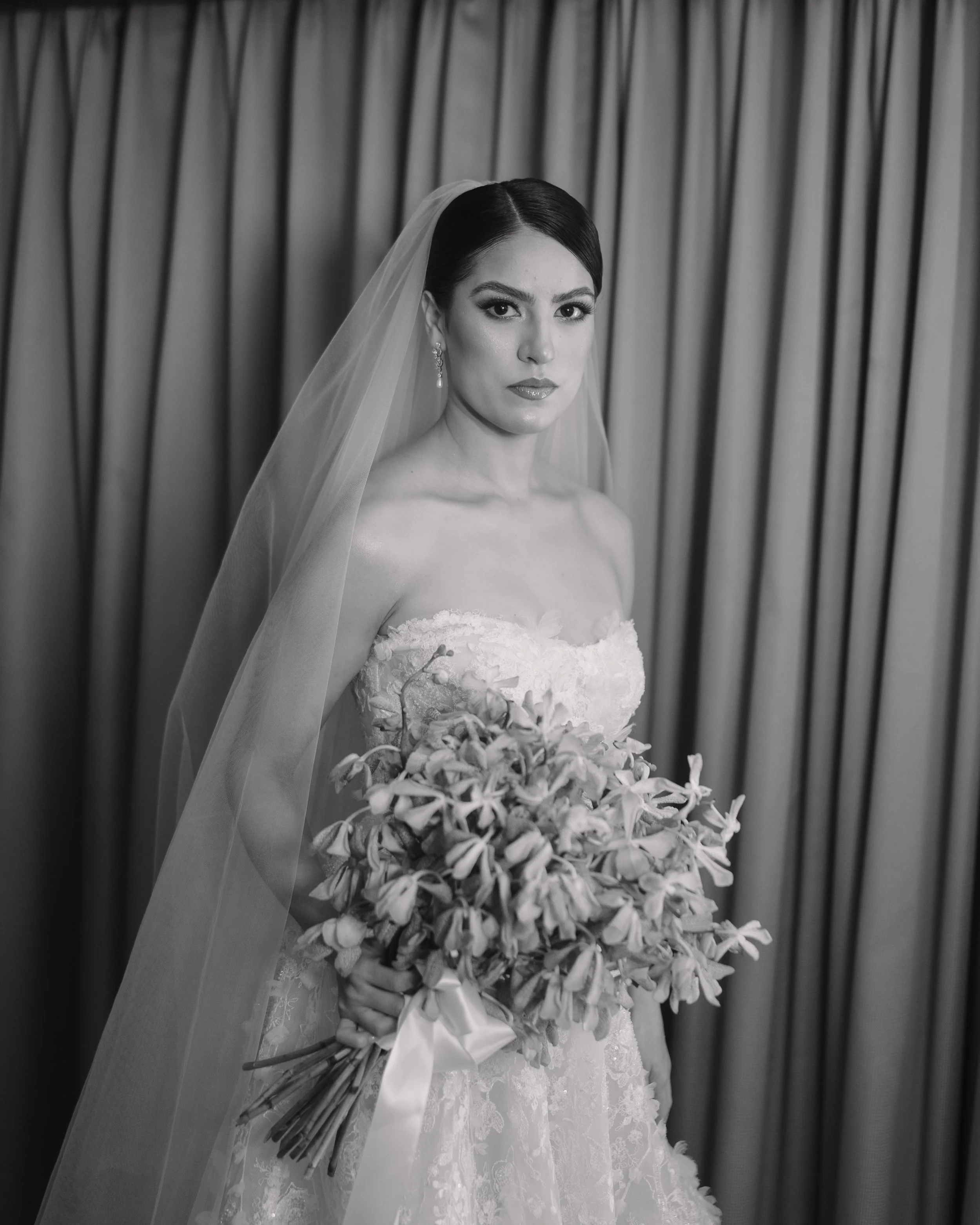 Black and white photo of a bride in a wedding dress holding a bouquet of flowers, standing in front of a curtain. Wedding Photography in Valle de Guadalupe, Ensenada, Baja California.