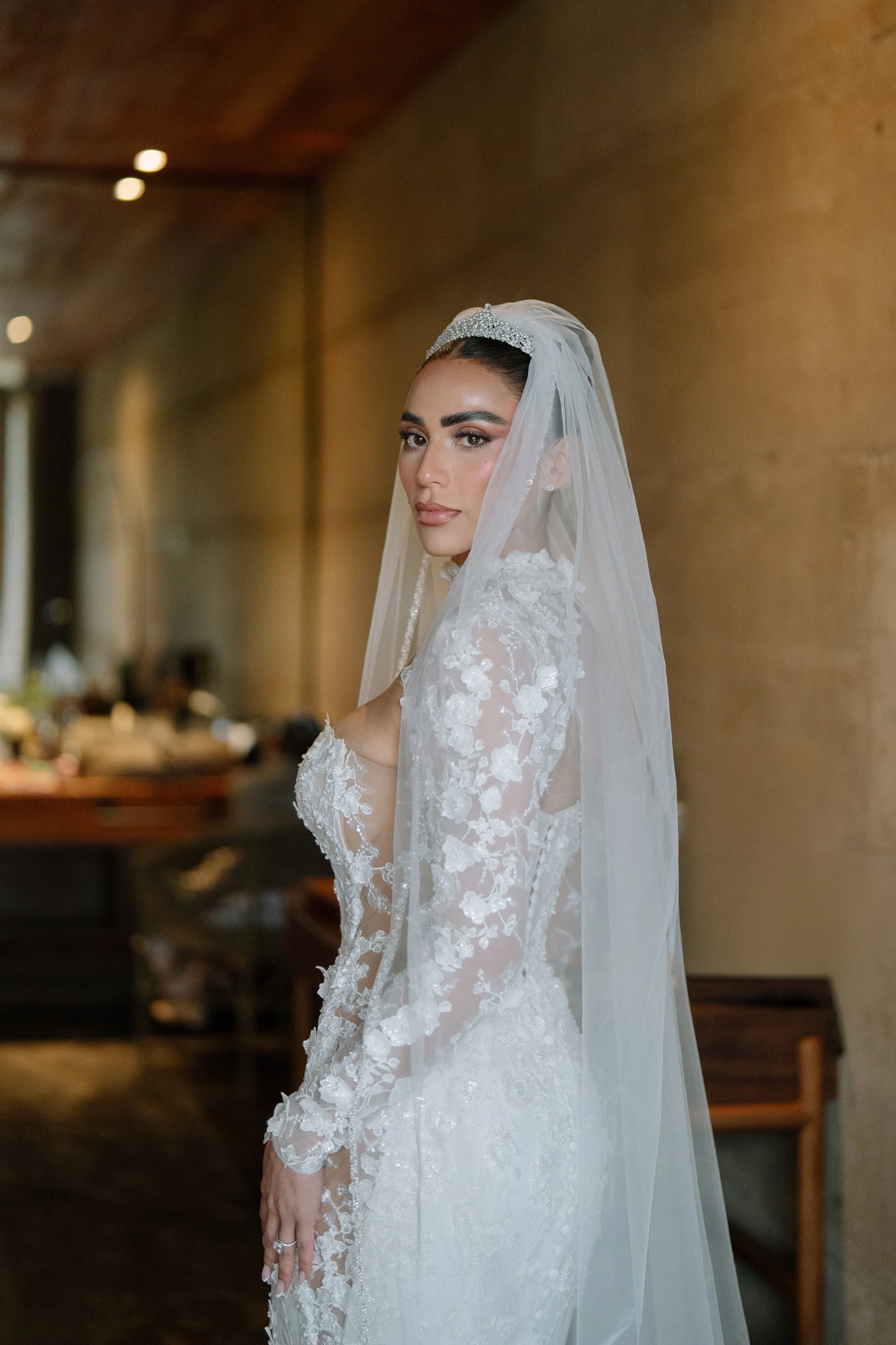 A bride in a white lace wedding dress and veil looking at the camera in a dimly lit room. Wedding Photography in Valle de Guadalupe, Ensenada, Baja California.