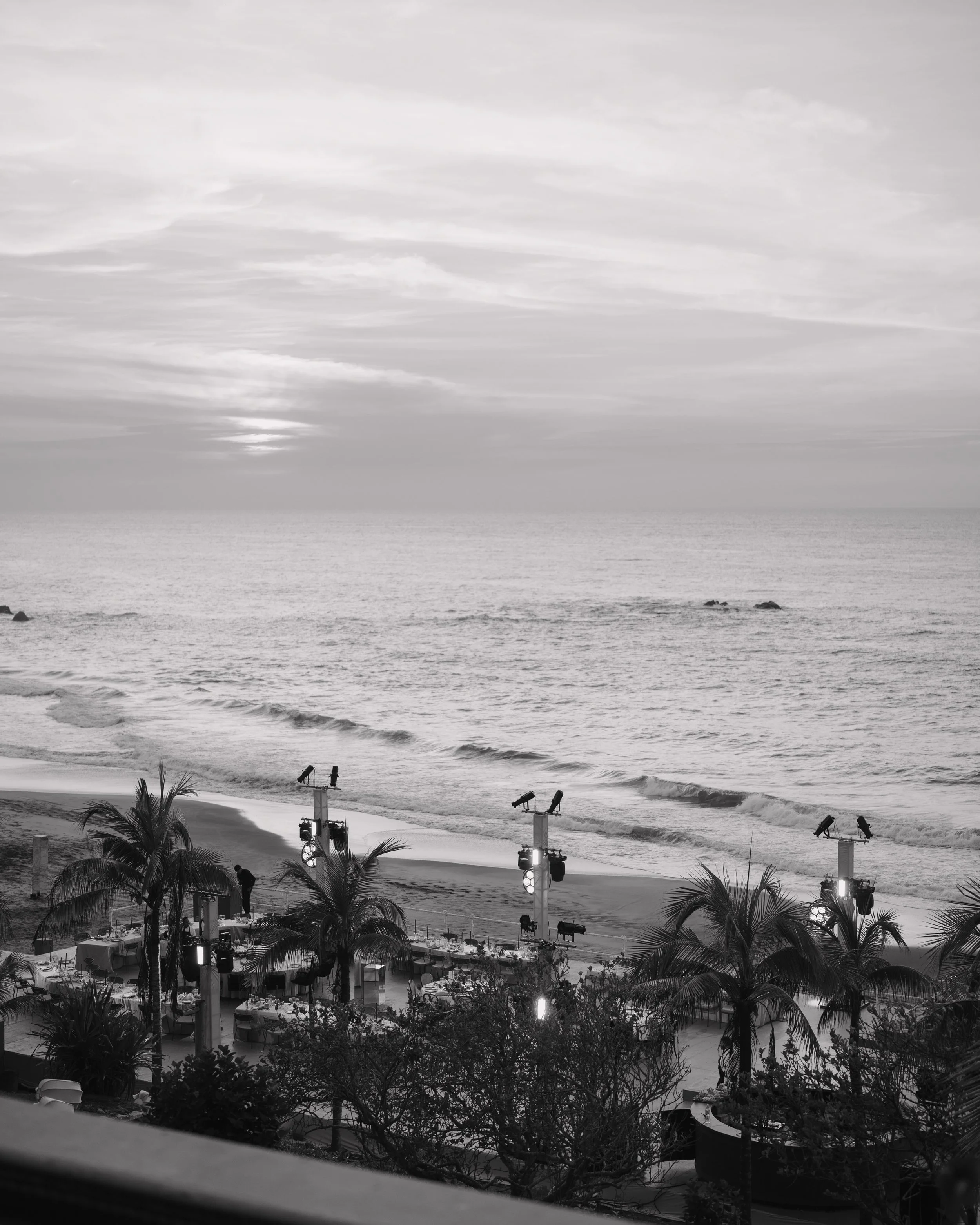 Black and white photograph of a beach scene with palm trees, stage lights, and a boat in the ocean during sunset. Wedding Photography in Valle de Guadalupe, Ensenada, Baja California.