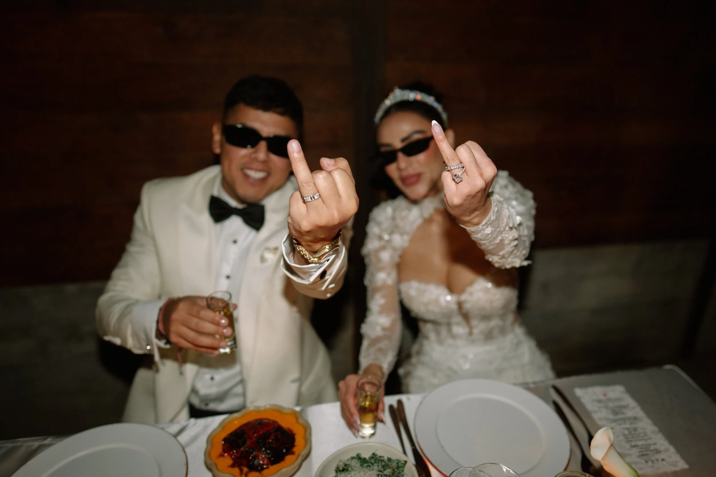 A newlywed couple, dressed in wedding attire, making a rude hand gesture while smiling and wearing sunglasses at their wedding reception. Wedding Photography in Valle de Guadalupe, Ensenada, Baja California.