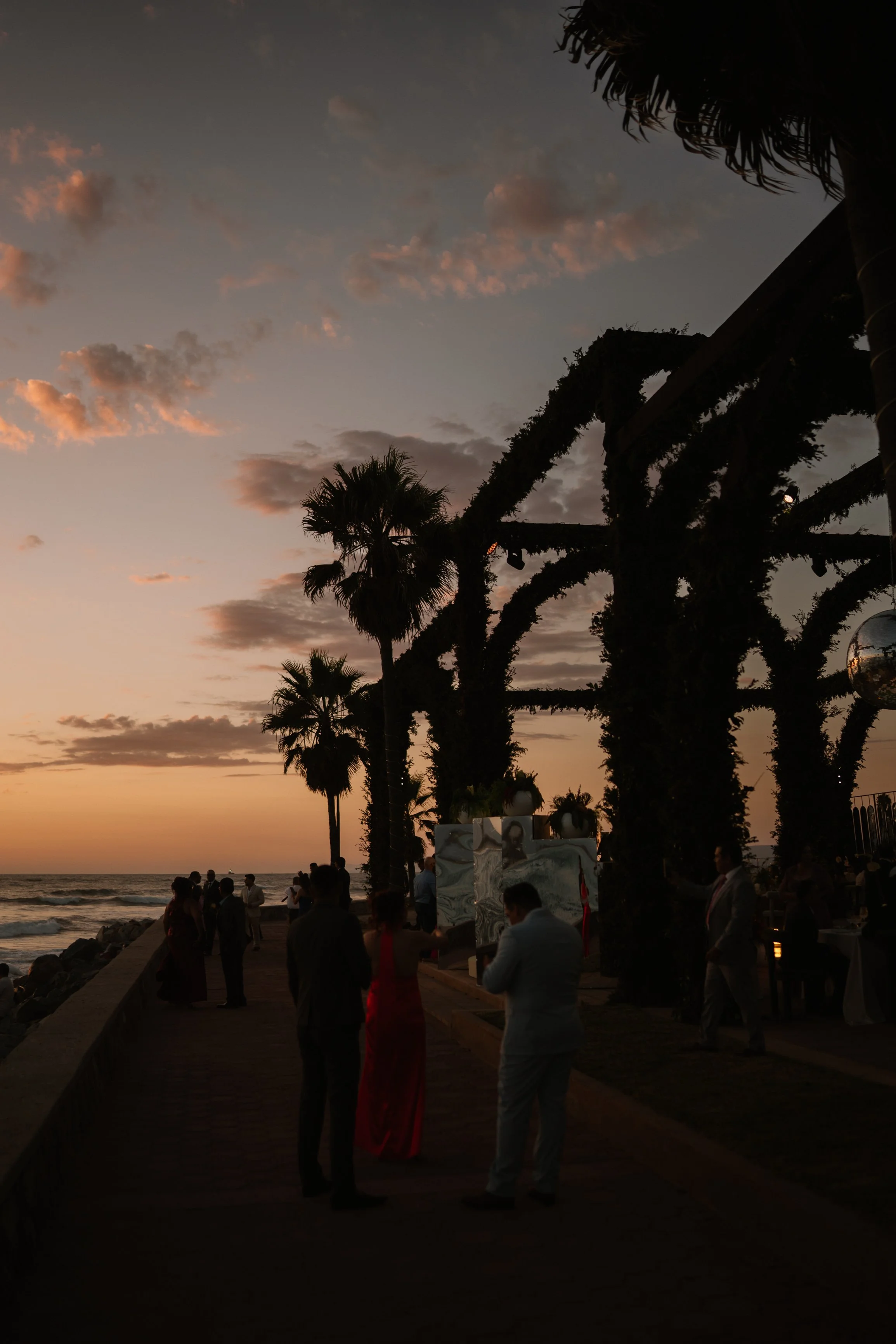 People dressed in formal attire gather on a beachside promenade at sunset, with palm trees and decorative structures under a partly cloudy sky. Wedding Photography in Valle de Guadalupe, Ensenada, Baja California.