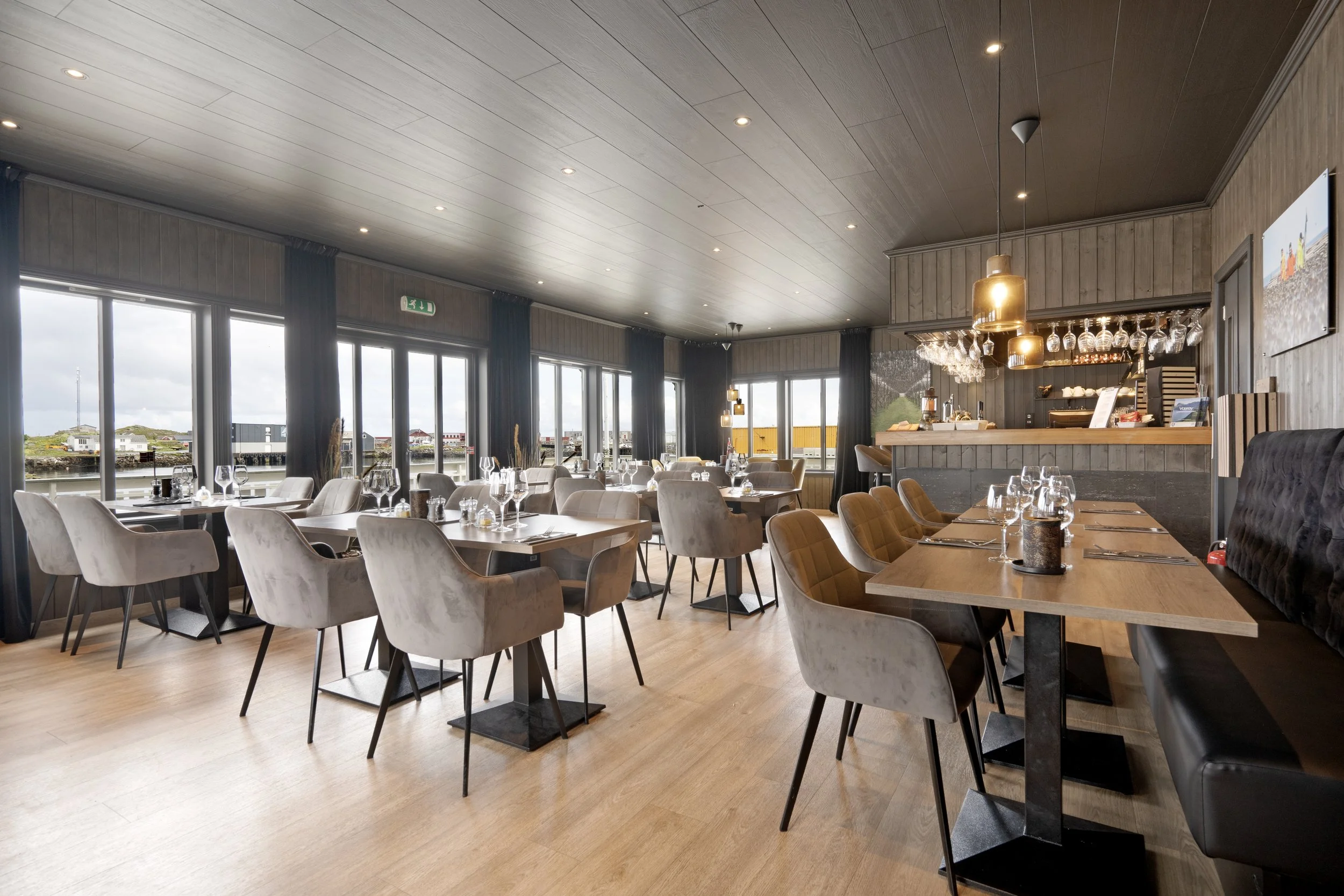 Elegant restaurant with large windows overlooking a waterfront, modern wooden ceiling, and a bar area with hanging glassware and wine glasses.