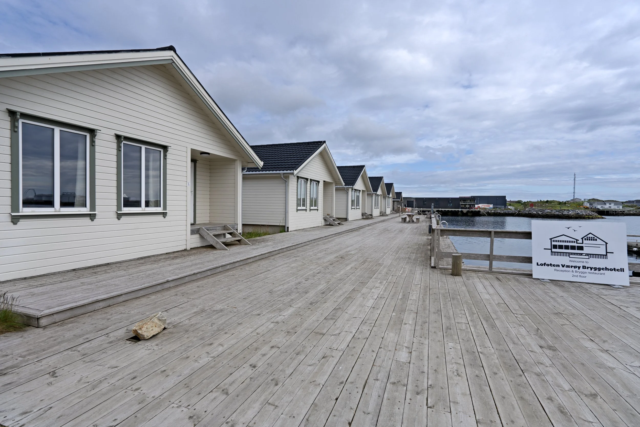 A row of white buildings with black roofs along a wooden boardwalk next to water, under a partly cloudy sky. A sign displays the name 'Lofoten Værøy Bryggesbotei'.