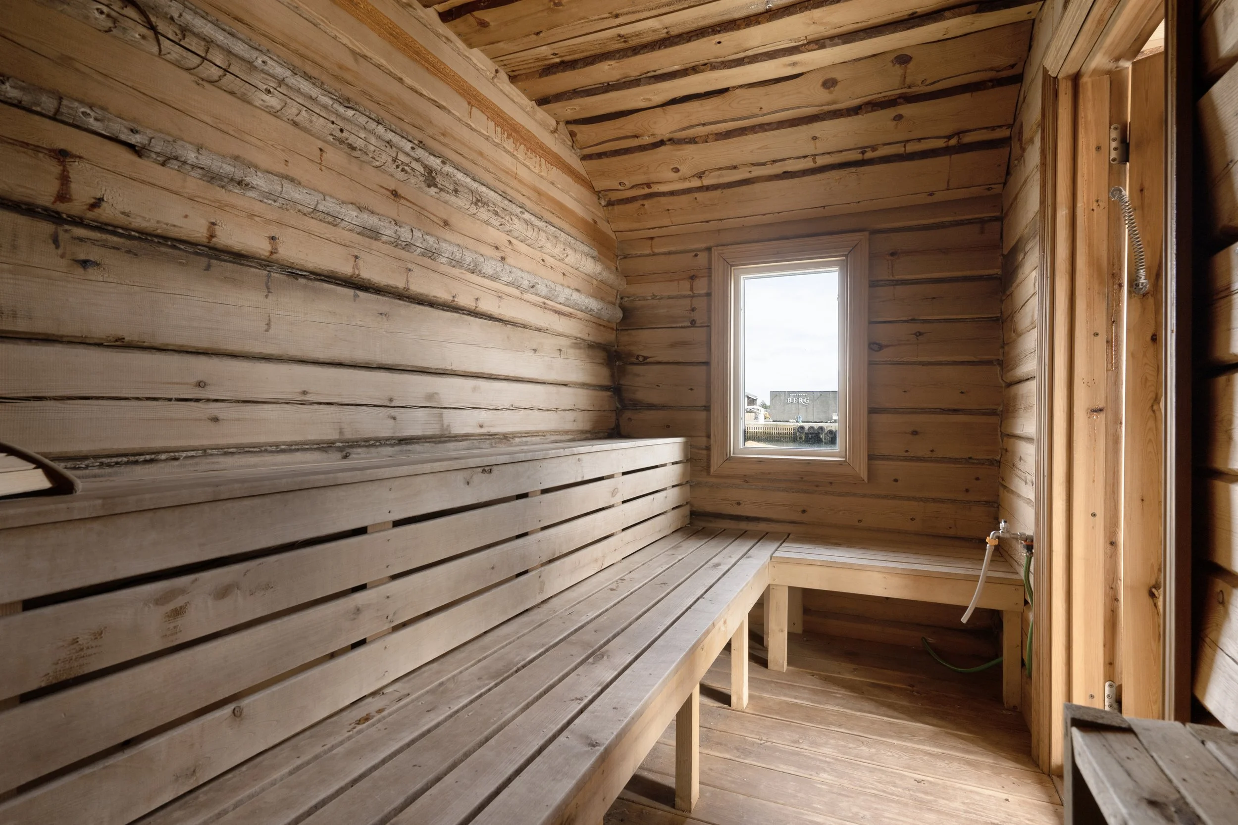 Interior of a small, unfinished wooden sauna with benches along the walls and a window showing a building in the background.