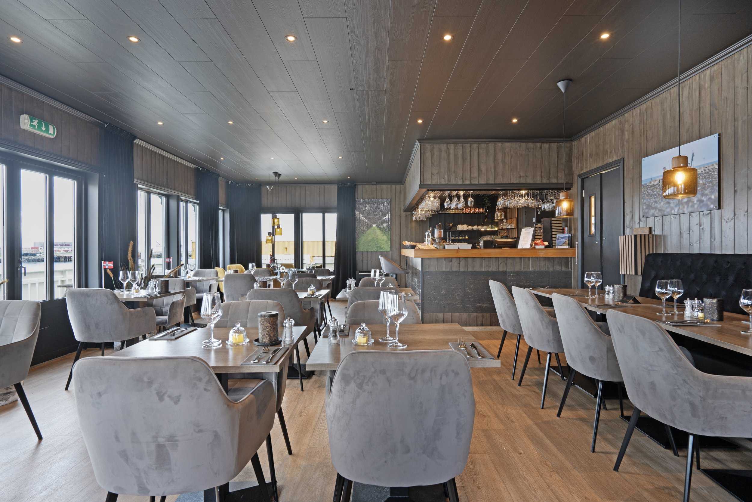A cozy restaurant with wooden-paneled walls and ceiling, large windows with blue curtains, and neatly set tables with wine glasses and candles, including a bar area with hanging glasses and a menu.