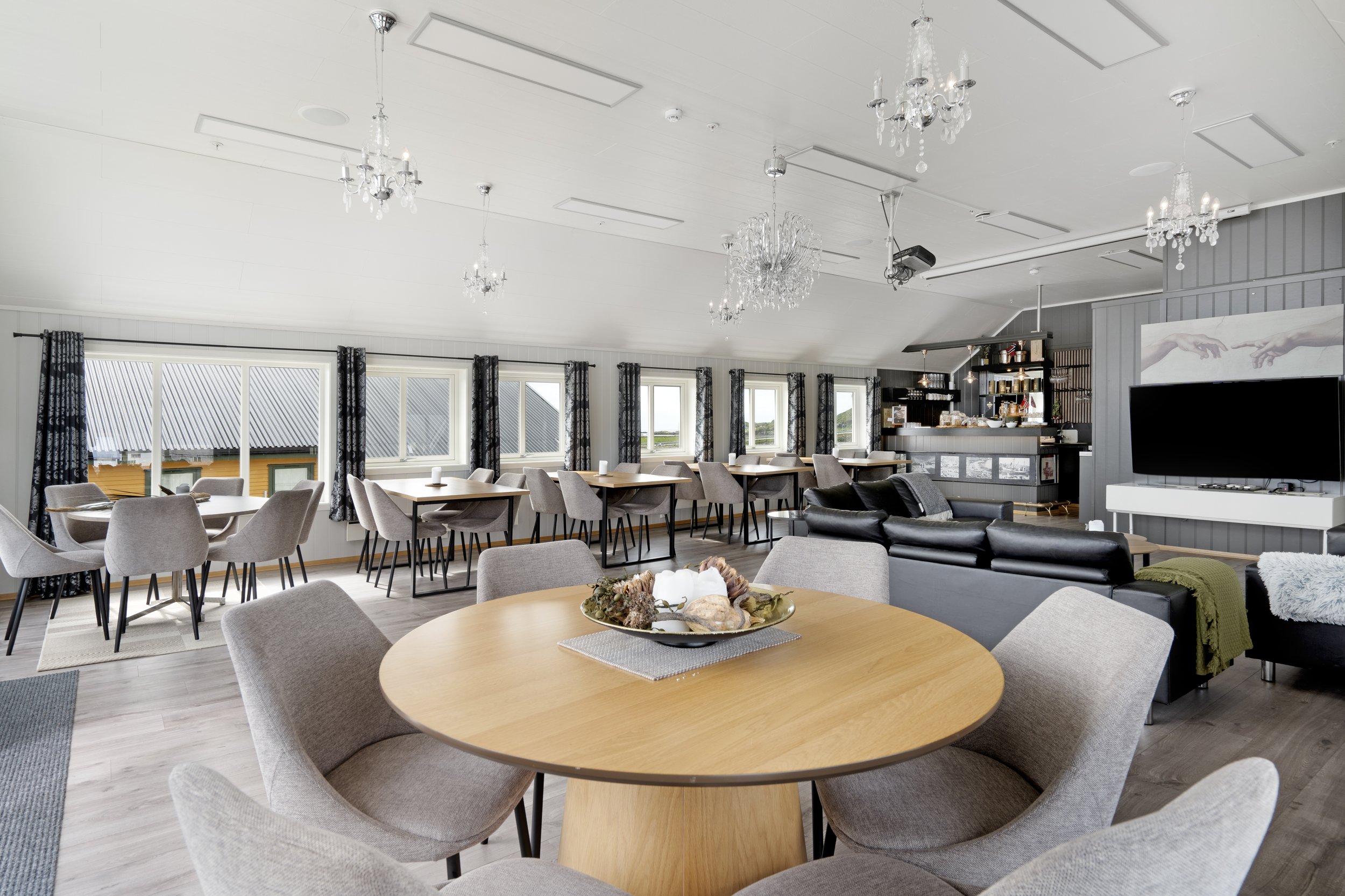A spacious dining and living area with multiple tables and chairs, large windows with patterned curtains, chandeliers hanging from the ceiling, and a modern TV on the wall with a painting of two hands reaching towards each other.