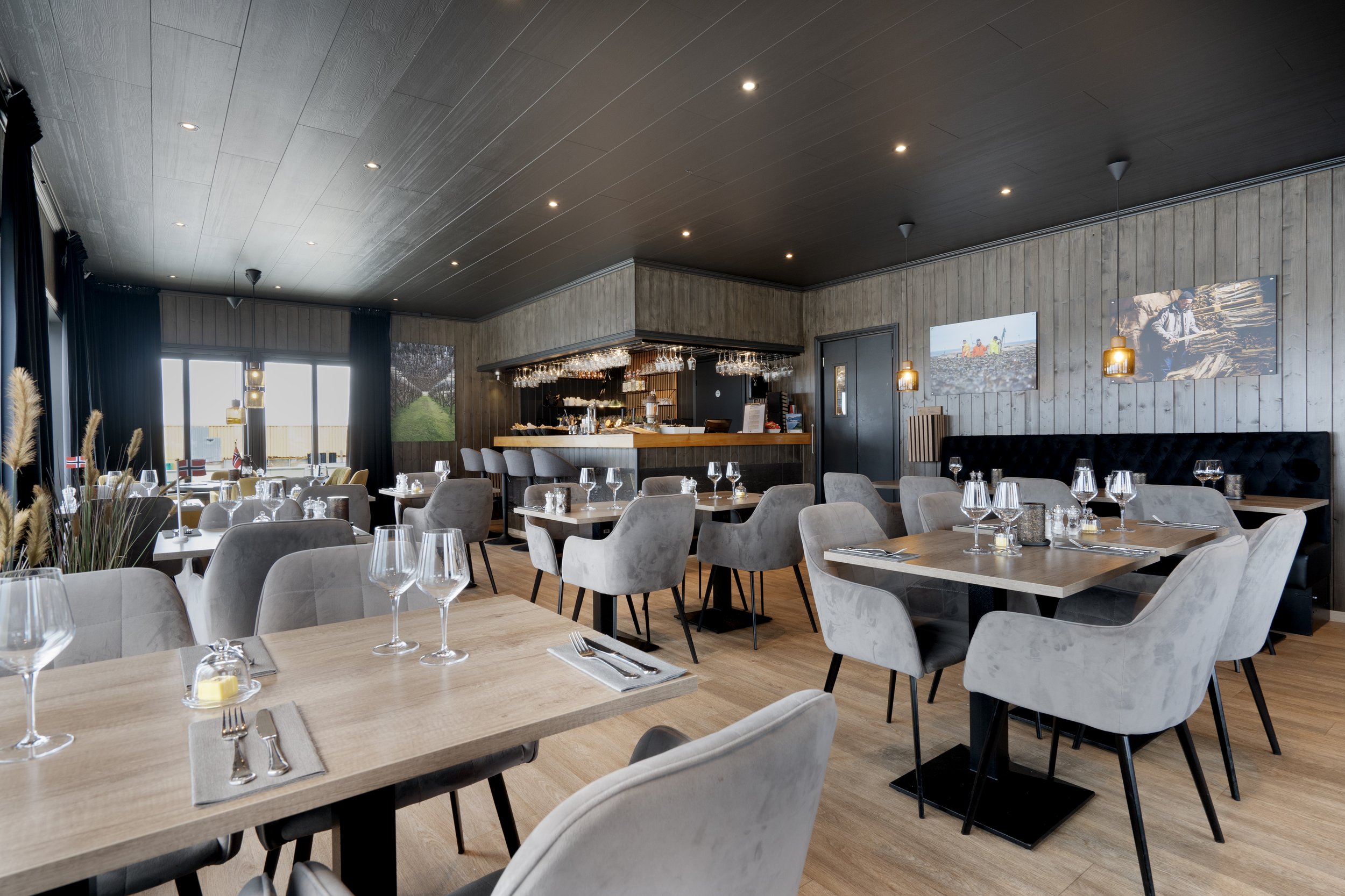 Elegant restaurant dining area with light wood tables set with wine glasses, silverware, and candles, gray upholstered chairs, large windows with dark curtains, wall-mounted photographs, and a bar area in the background.