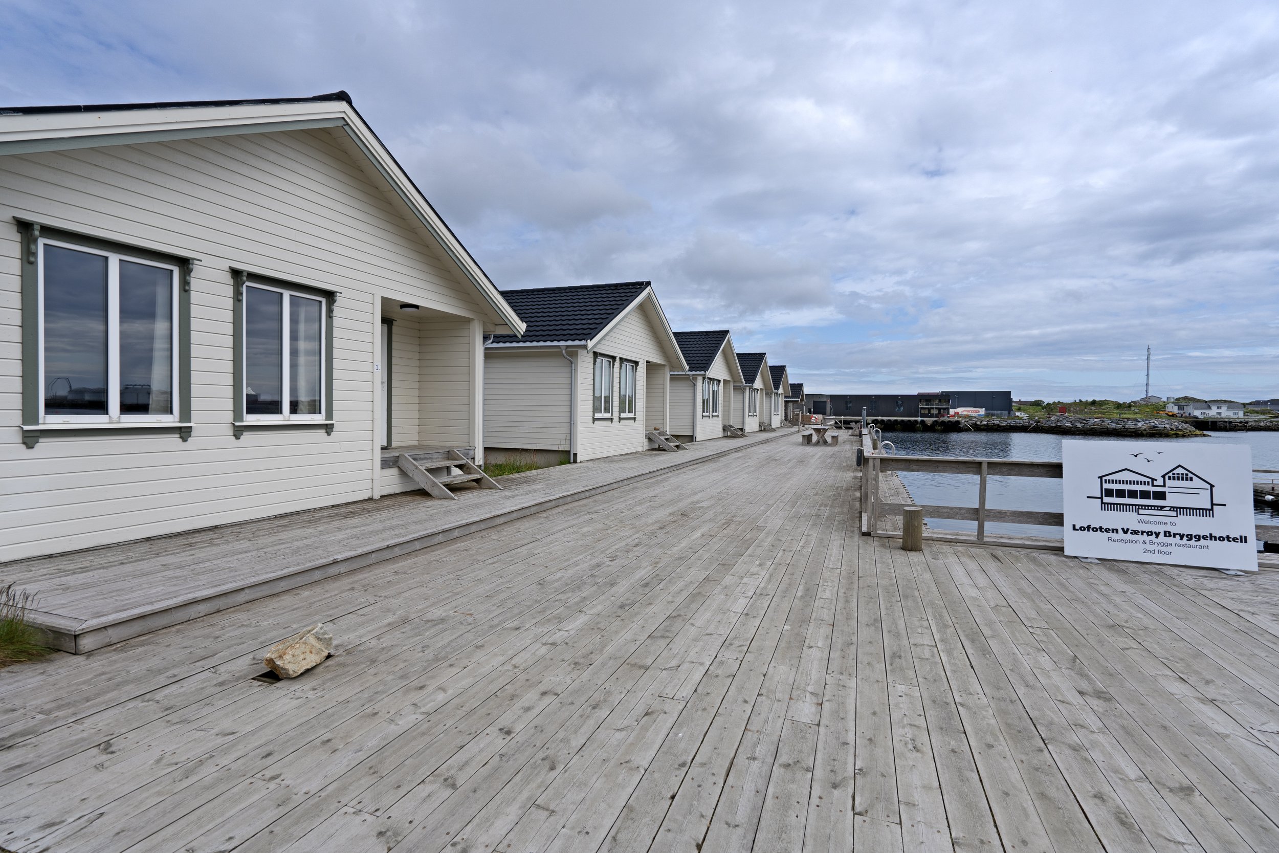 White wooden houses with dark roofs along a water body under cloudy sky, with a sign that reads "Lofoten Værøy Brygghotel" and indicates renovation and dining services.
