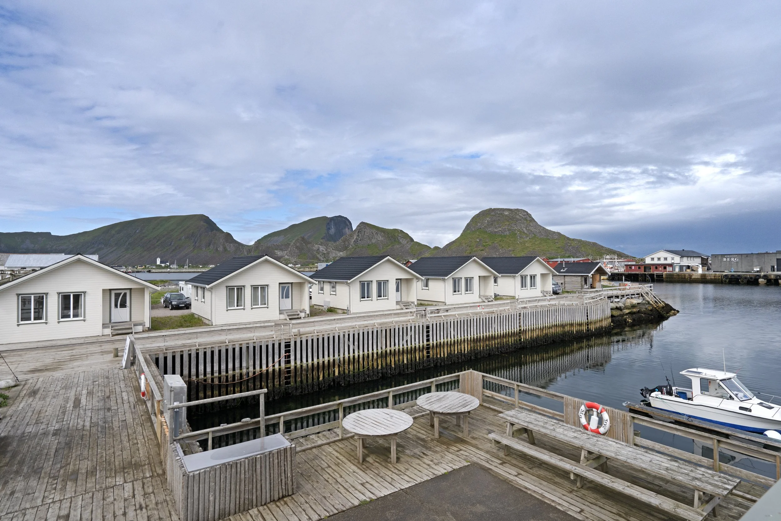 White cottages with black roofs lined along a wooden pier by the water, with a small boat docked nearby, mountains in the background, under a partly cloudy sky.