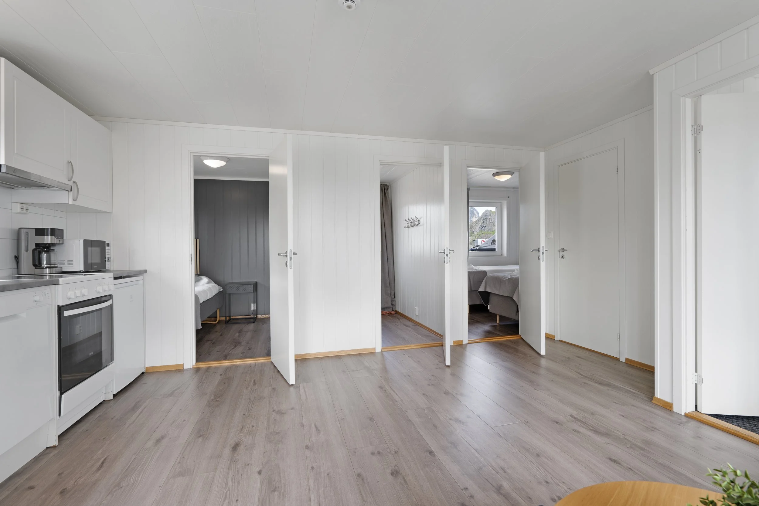 Interior of a house with white walls, a kitchen area on the left, and a hallway with three open doors leading to bedrooms. Wooden flooring and a ceiling light fixture are visible.