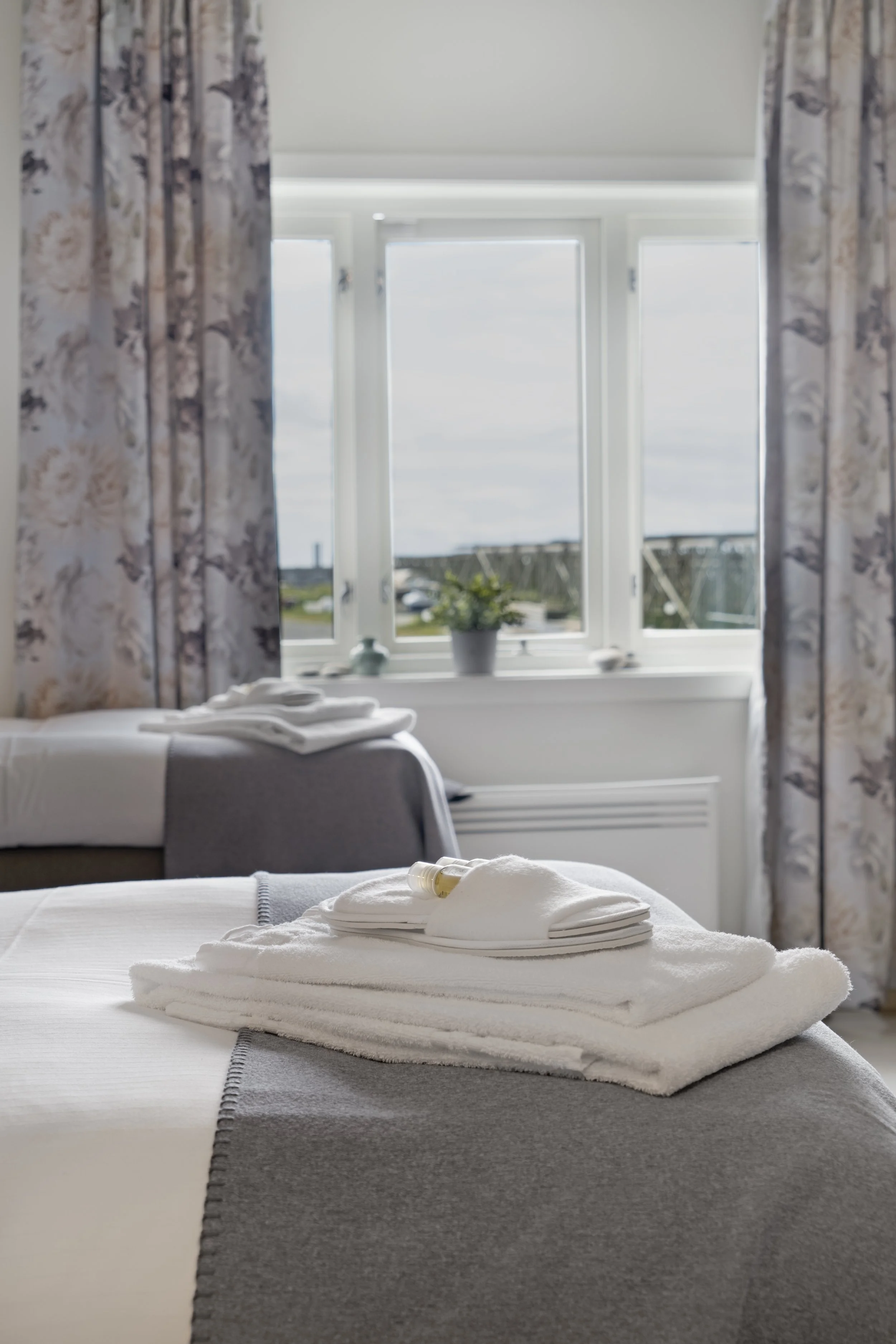 A neatly made bed with white linens, on which are folded towels and a small bottle, in a bright room with a large window and floral curtains.