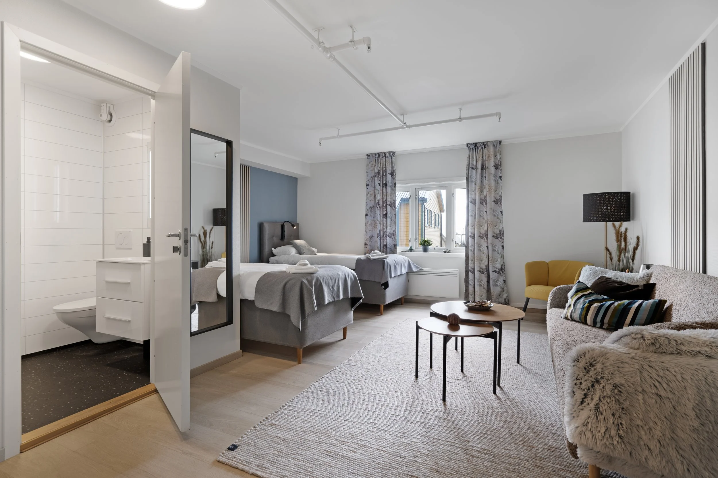A modern, minimalist apartment living space with an open view into a bedroom and bathroom. The room features two beds with gray bedding, a small round coffee table, a cozy sofa with a throw blanket, and a yellow armchair. Large window with floral curtains lets in natural light, and indoor plants decorate the window sill. The bathroom, visible through an open side door, has white tiled walls and a toilet. The overall decor is neutral with subtle accents and wooden flooring.
