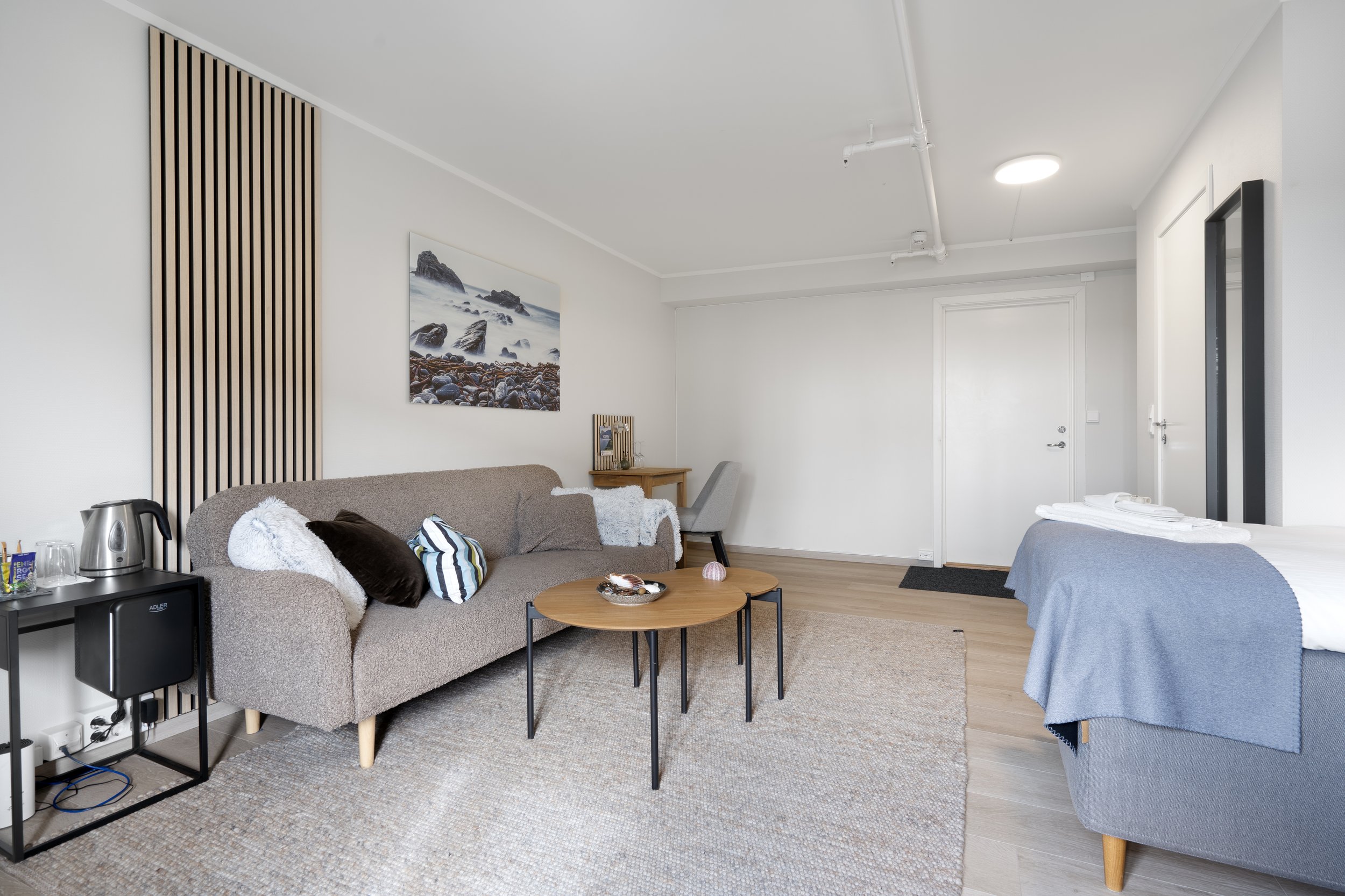 Interior of a modern hotel room featuring a seating area with a beige sofa, a round coffee table, and decorative pillows, a bed with a blue blanket, a wooden desk with a chair, and wall art of a rocky shoreline. There is a white door and a wall-mounted television.