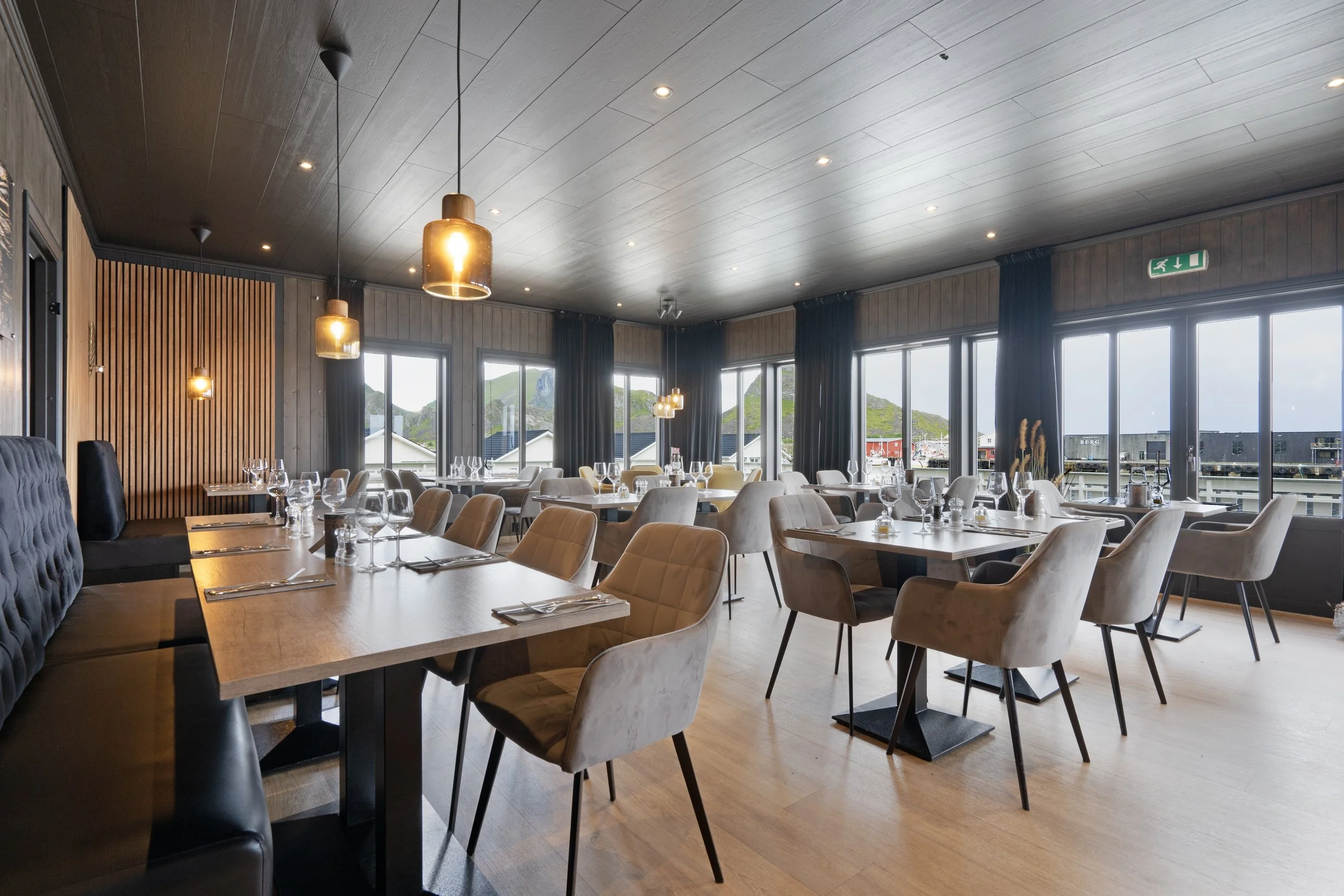 Interior of a modern restaurant with large windows, wooden paneling, hanging pendant lights, and tables set with glassware and napkins.