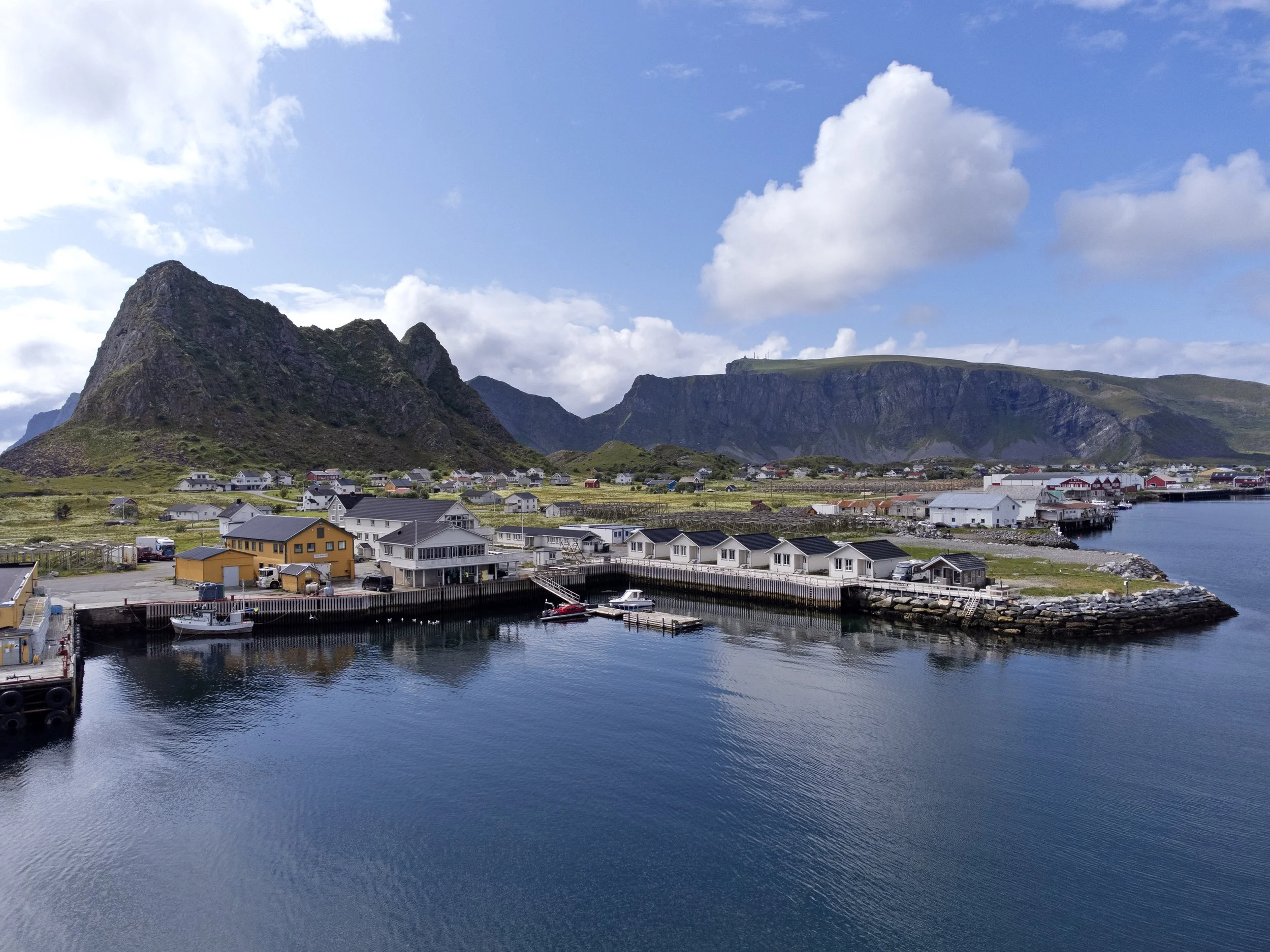 A coastal village at the base of green mountains with a harbor of docked boats and houses, under a partly cloudy sky.