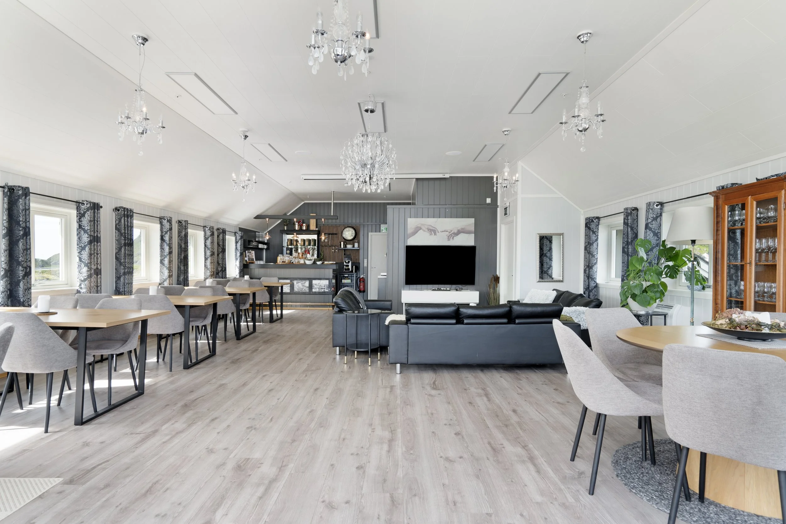 Interior of a spacious, modern dining and lounge area with large windows, chandeliers, gray and white decor, and various seating arrangements.