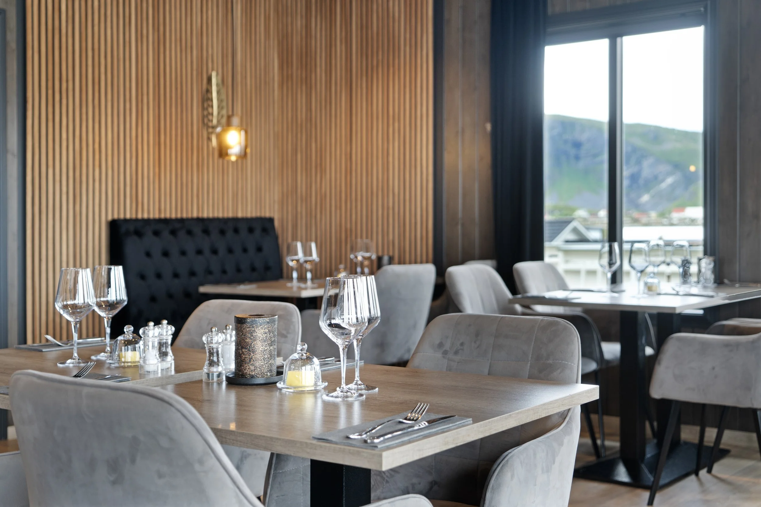 Elegant dining room with wooden accent wall, large window showing a view of hills, tables set with wine glasses, candles, and silverware, and upholstered chairs.