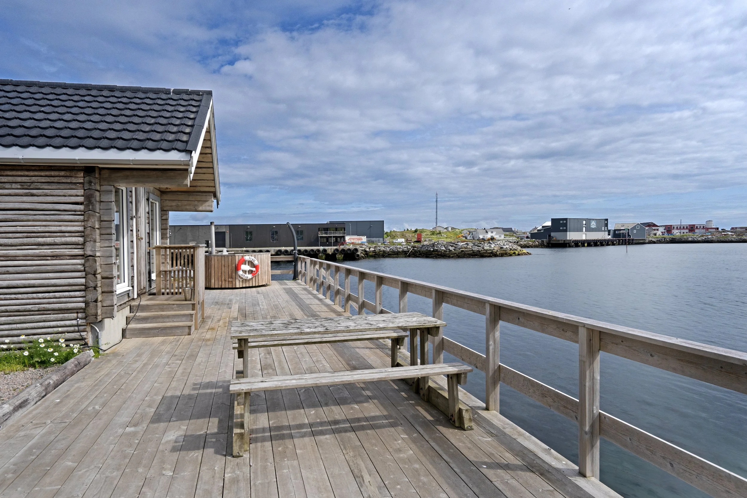 A wooden deck with benches and a railing looking out over calm water. A small wooden building is on the left, with a life preserver hanging on the railing. In the background, there are industrial and residential buildings, and a tall antenna against a partly cloudy sky.