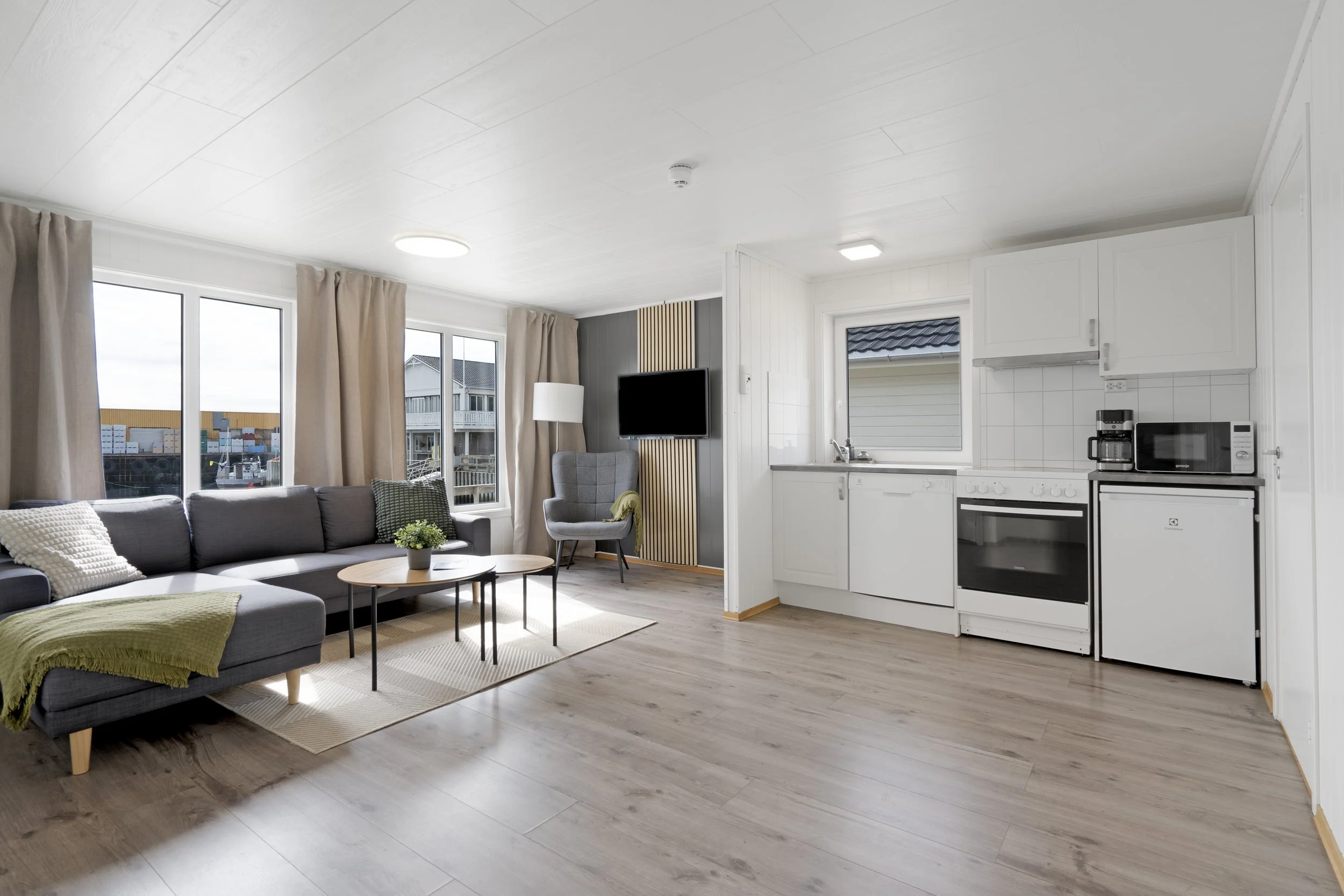 Open-plan living room and kitchen area with large windows, beige curtains, gray sofa with cushions, round coffee table, kitchen with white cabinets, stove, microwave, coffee maker, and a window with a view of neighboring buildings.