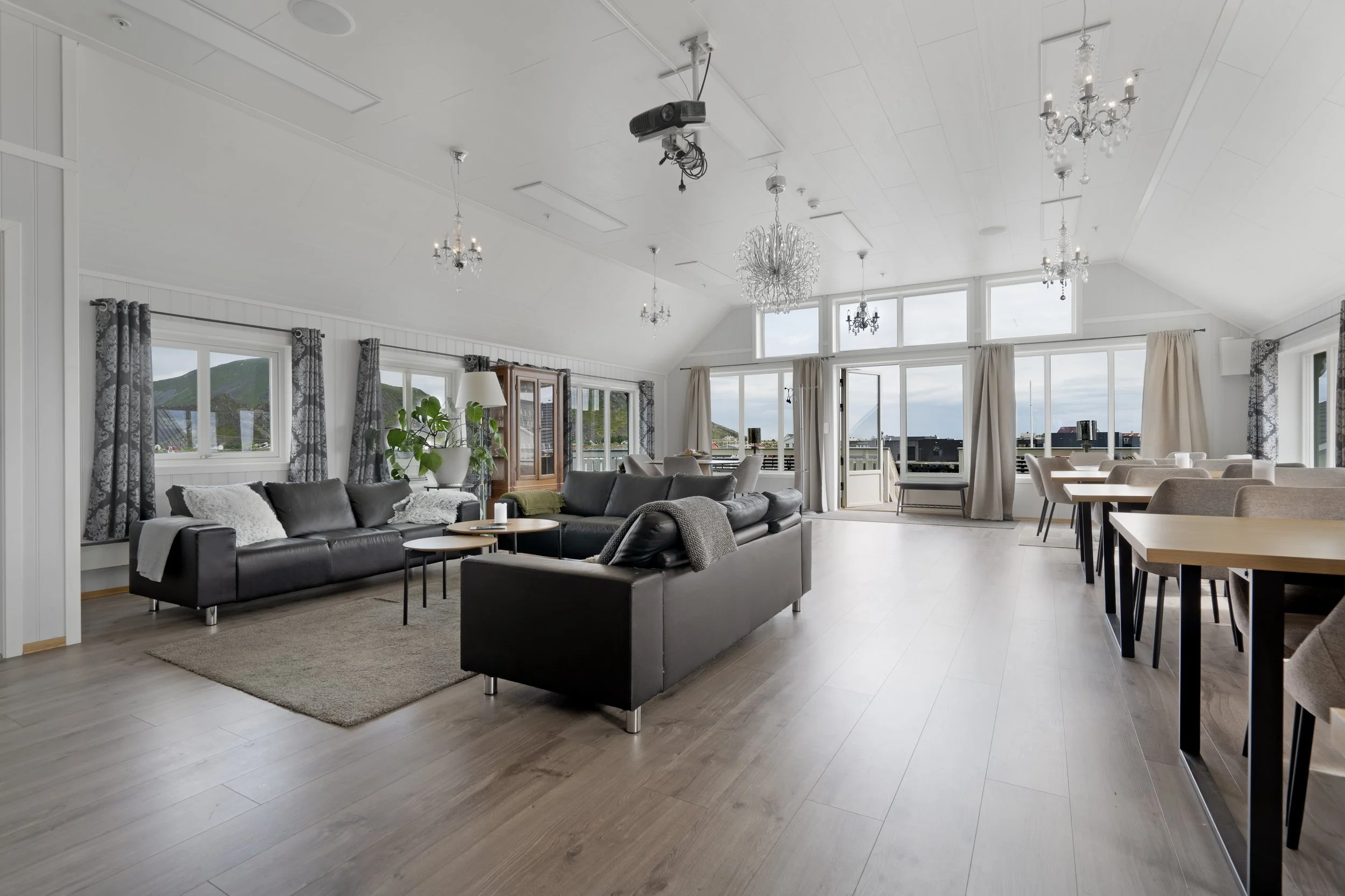 A spacious living room with large windows, white walls, and wooden flooring. It features black leather sofas, a gray carpet, a wooden dining table with beige chairs, and decorative chandeliers hanging from the white ceiling. There are curtains on the