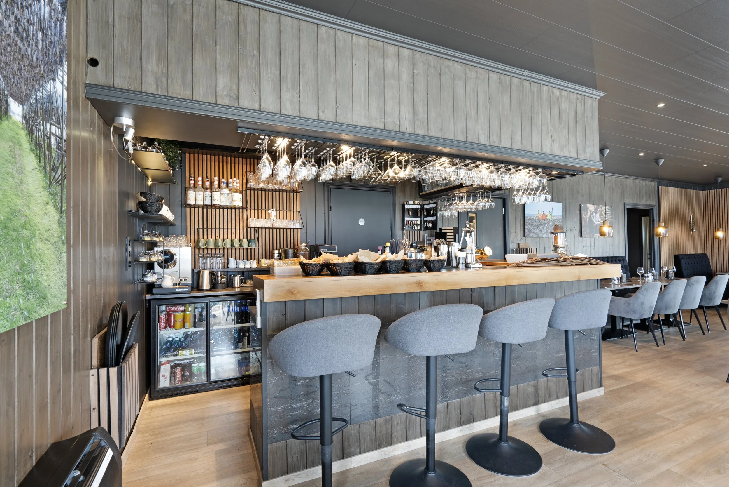 Interior view of a modern bar or restaurant with high-top tables and chairs, pendant lighting, glasses hanging upside down above the bar, and a small refrigerator with drinks.