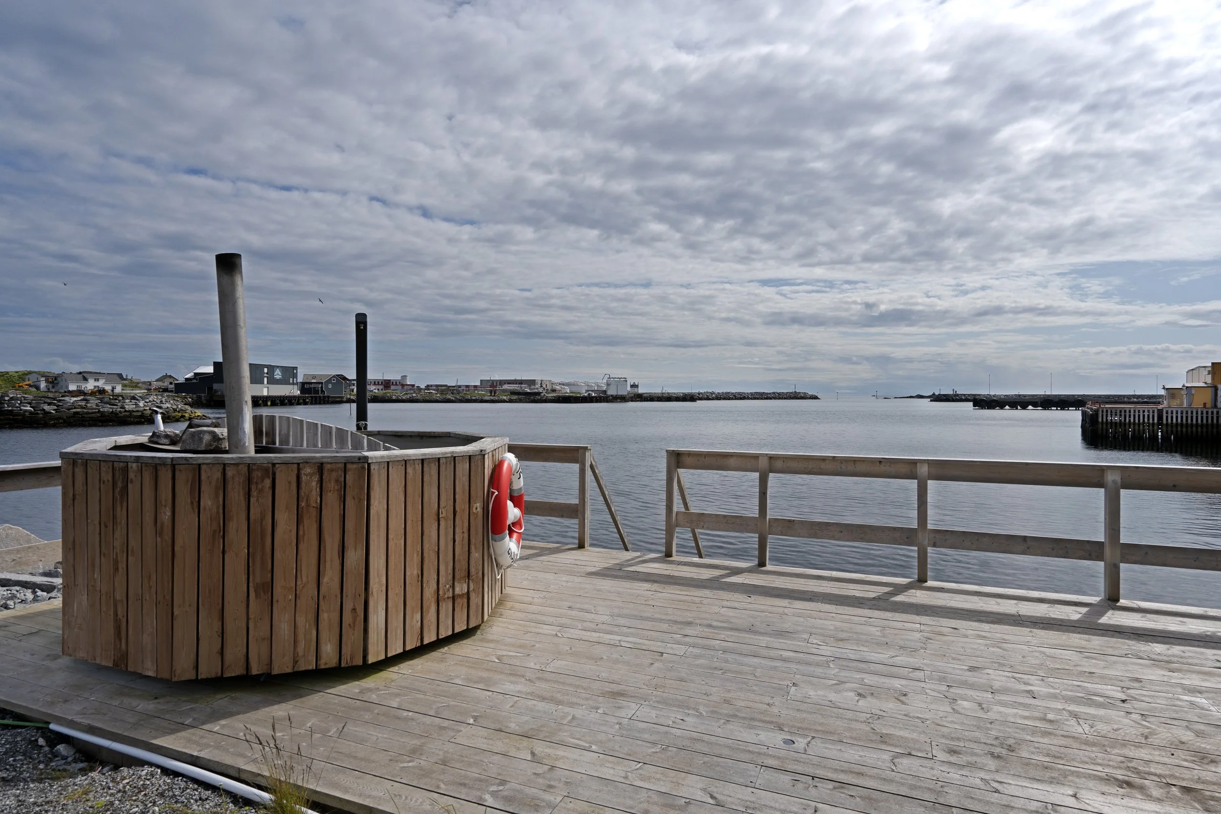 Wooden deck by the water with a life preserver hanging on the side. In the background, there are boats, buildings, and a cloudy sky over a harbor.