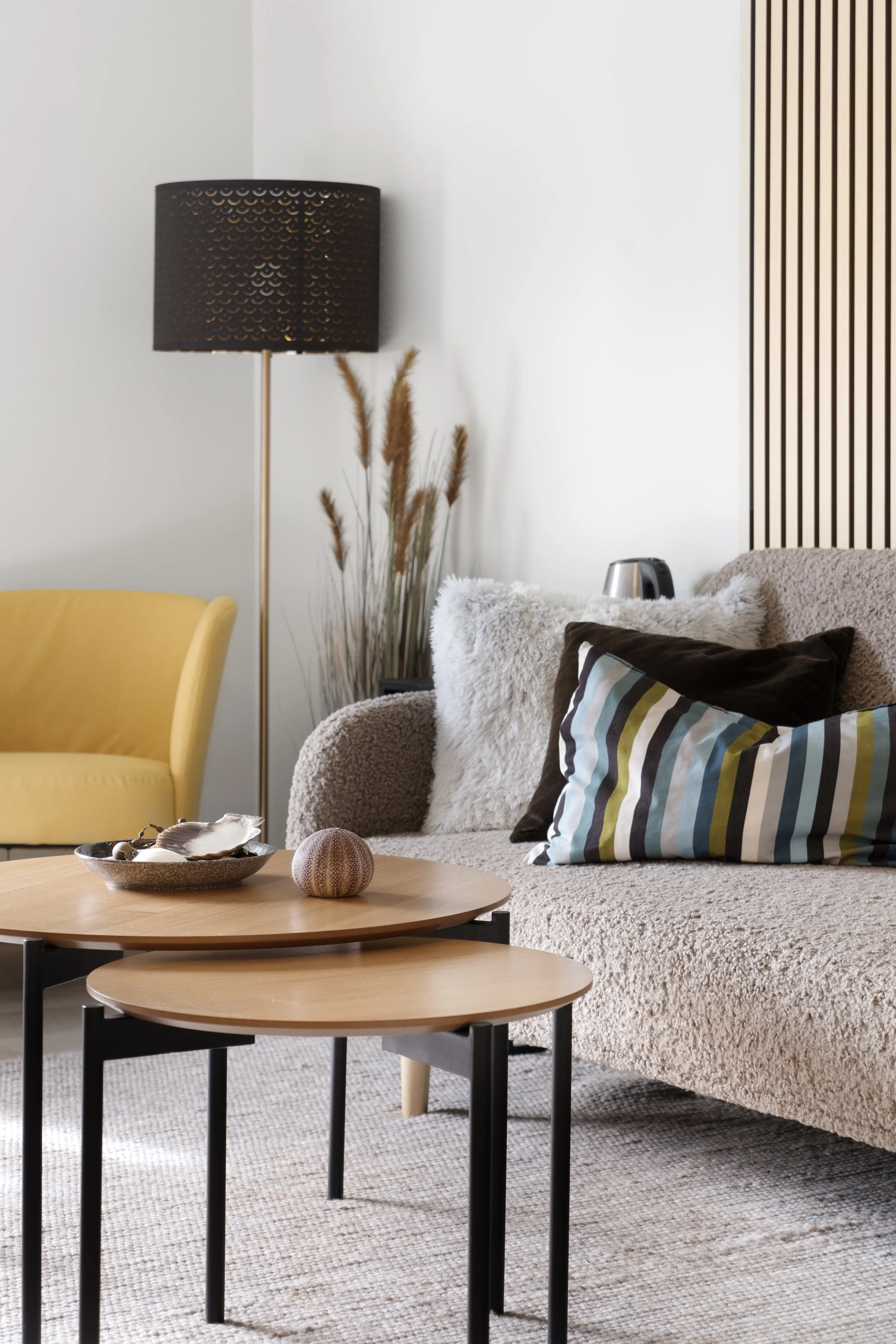 Living room with a beige sofa with various pillows, a round wooden coffee table with decorative objects, a yellow armchair, a black floor lamp, tall dried plants, and a wooden wall accent.