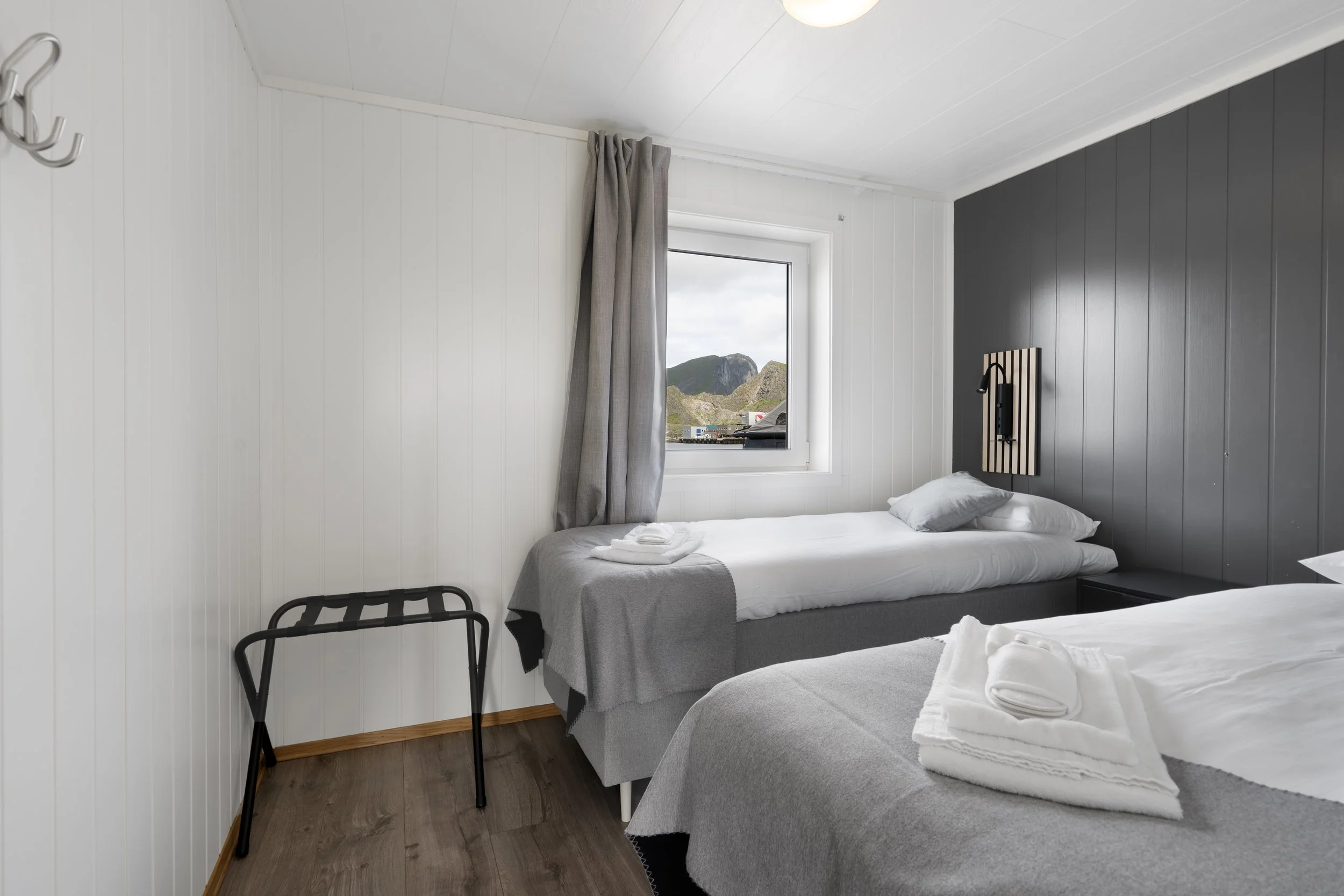 A bedroom with white and gray walls, a window showing mountainous landscape, two beds with white linens and folded towels, a black chair, and a nightstand.