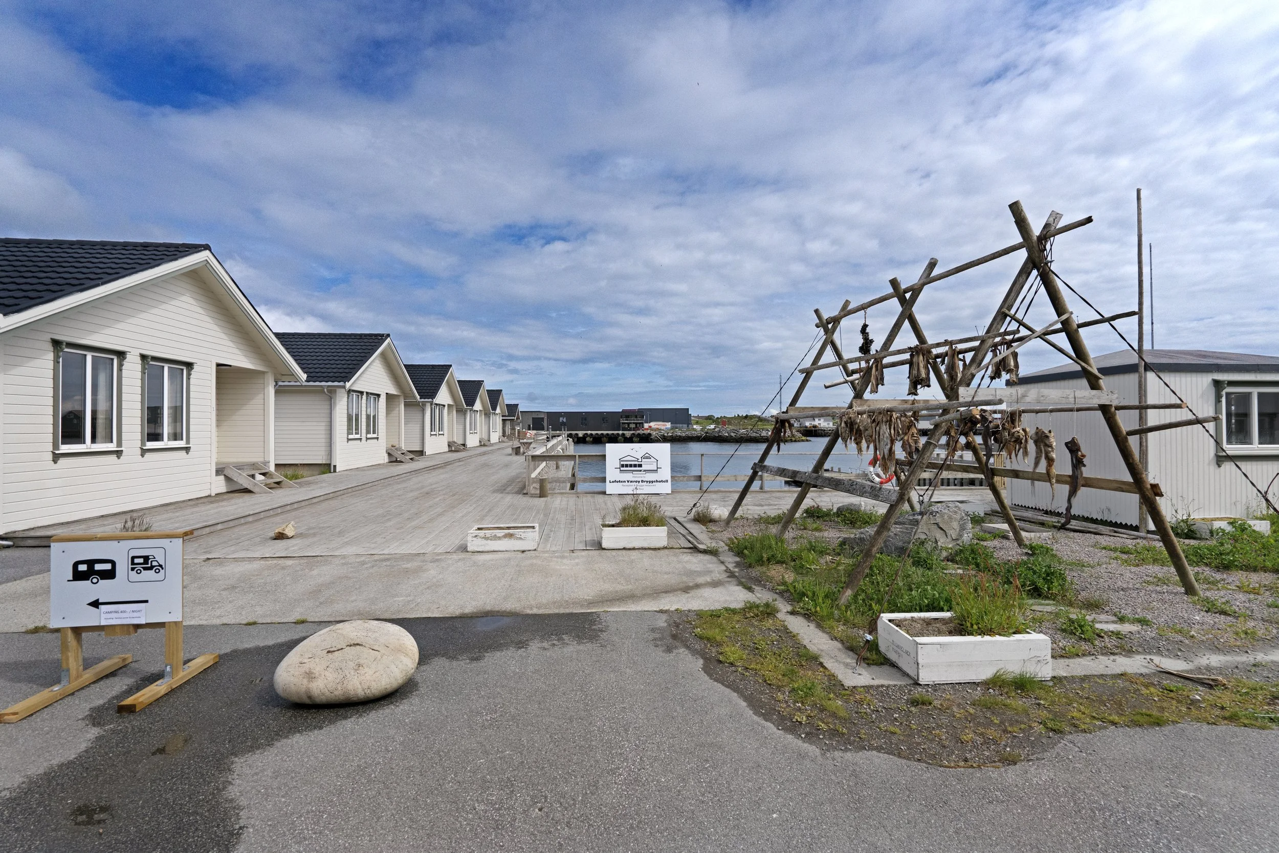 A row of white houses with black roofs along a dock, a wooden sculpture resembling a ship, and signboards indicating camping and RV parking, under a cloudy sky.