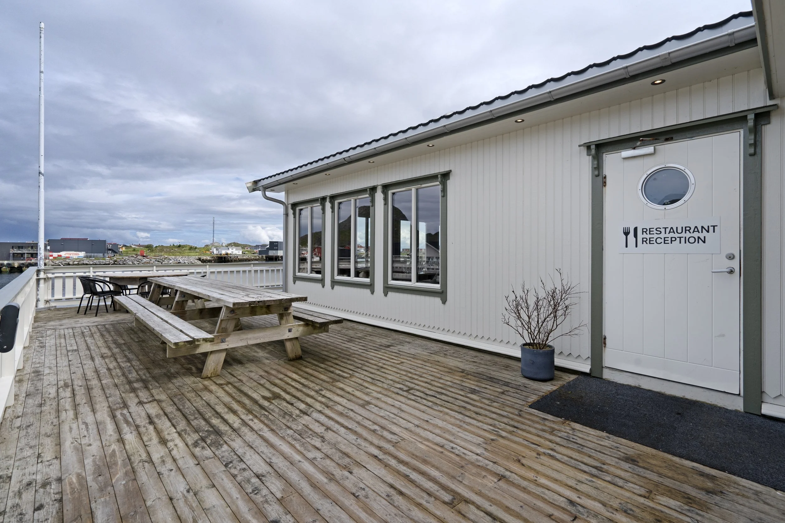 An outdoor wooden deck with a picnic table and two chairs, adjacent to a white building featuring a sign that reads 'Restaurant Reception.' The sky is cloudy, and there are buildings and a distant landscape visible beyond the deck.