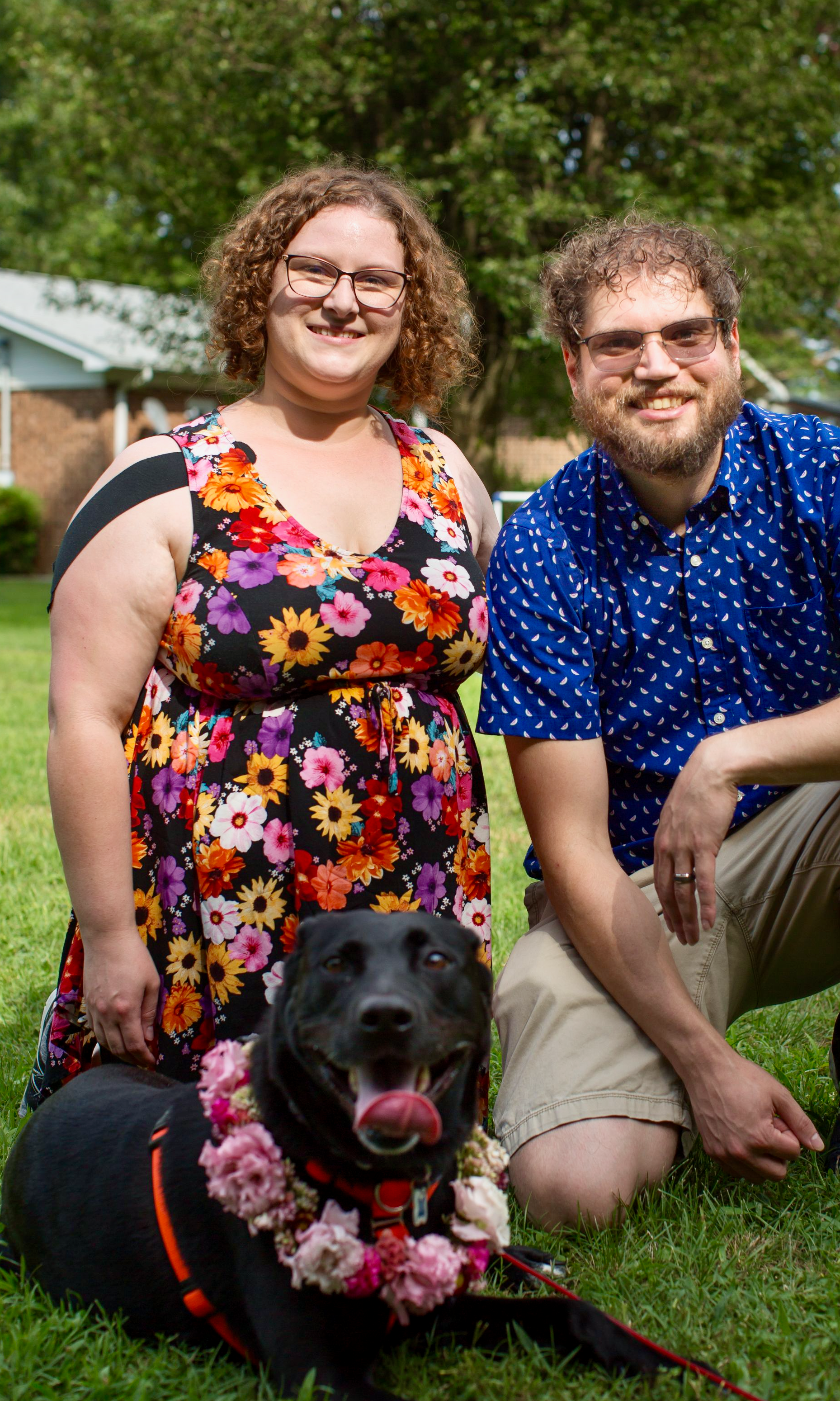 A smiling woman with curly hair and glasses wearing a colorful floral dress a smiling man with glasses and a beard wearing a blue patterned shirt, and a black dog with a pink floral lei on its neck, all outdoors on a grassy lawn with trees and a house in the background.