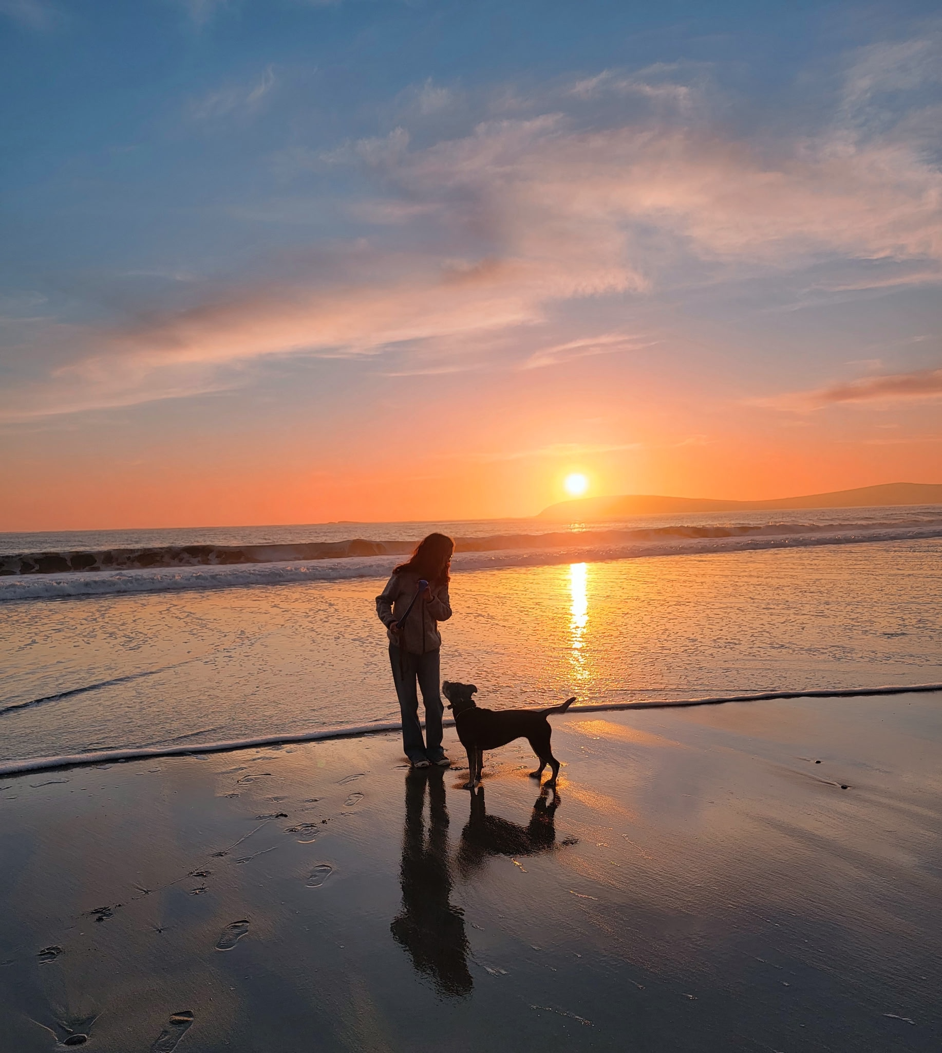 A woman walking her dog on the beach during sunset, with the reflected sunlight on the wet sand and a colorful sky, calm sea waves in the background.