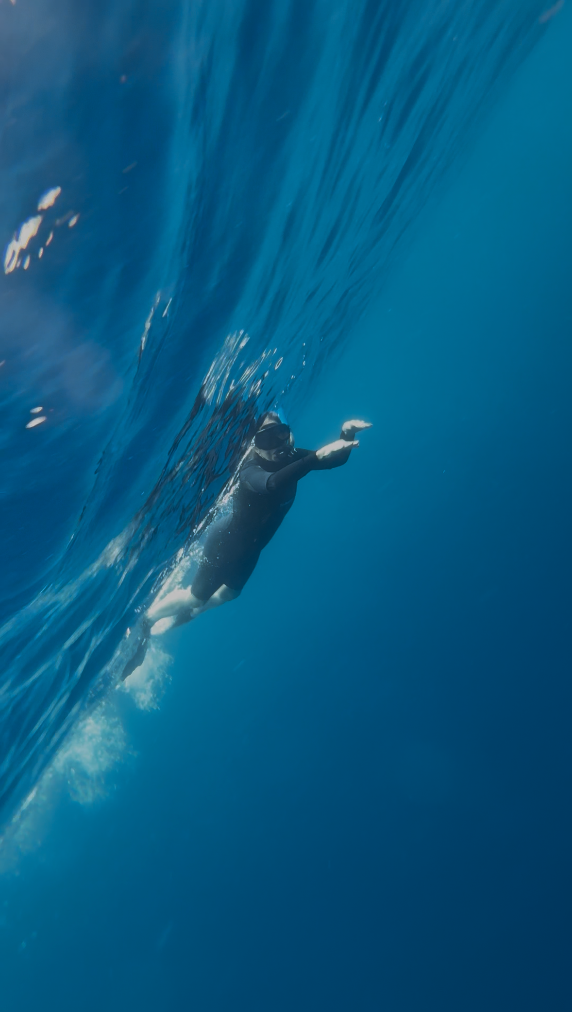 Person in black swimwear and snorkeling gear swimming under the water with a wave above.
