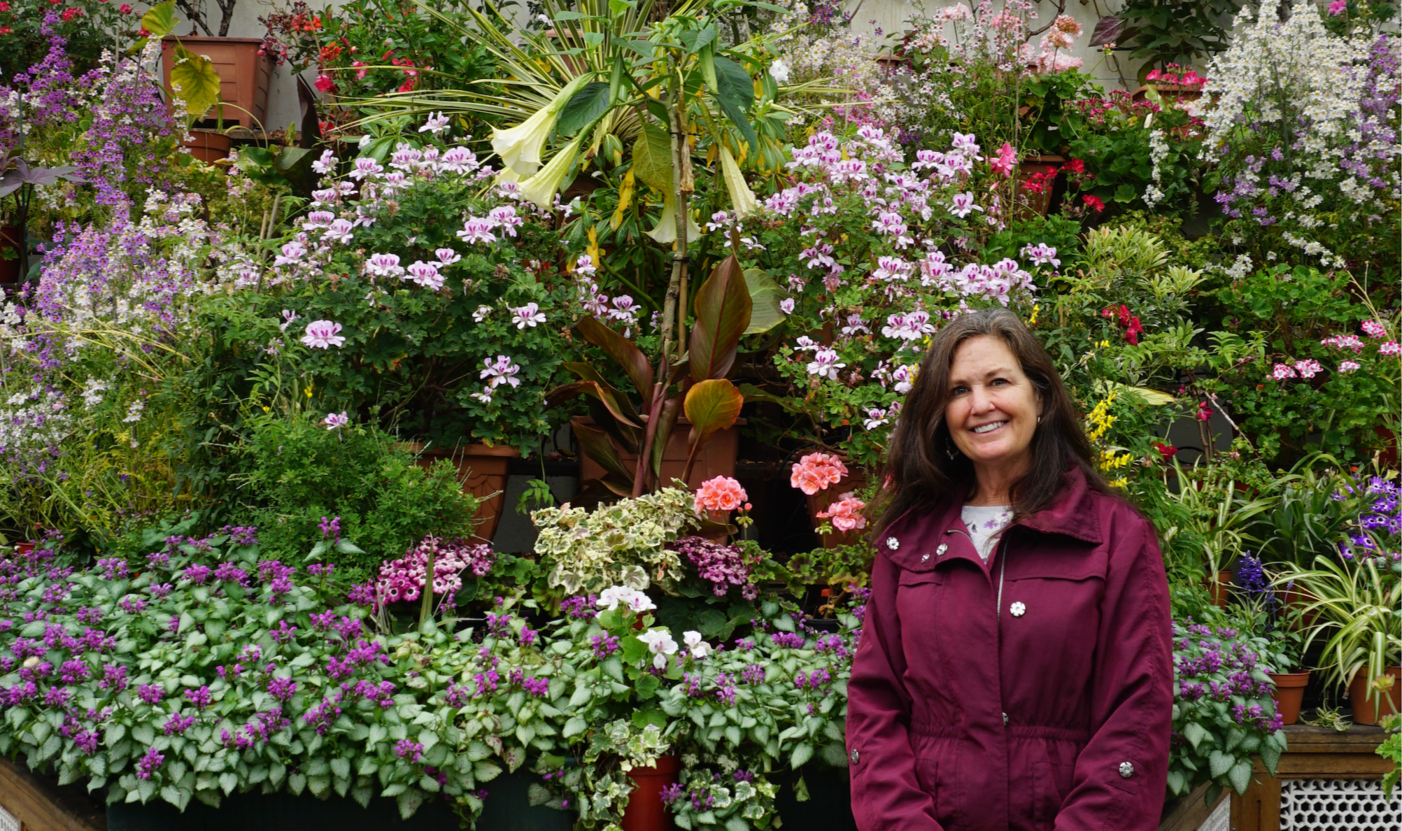 A woman with long brown hair smiling in front of a lush display of various colorful flowers, including pink, purple, and white blooms, in a garden or plant shop.