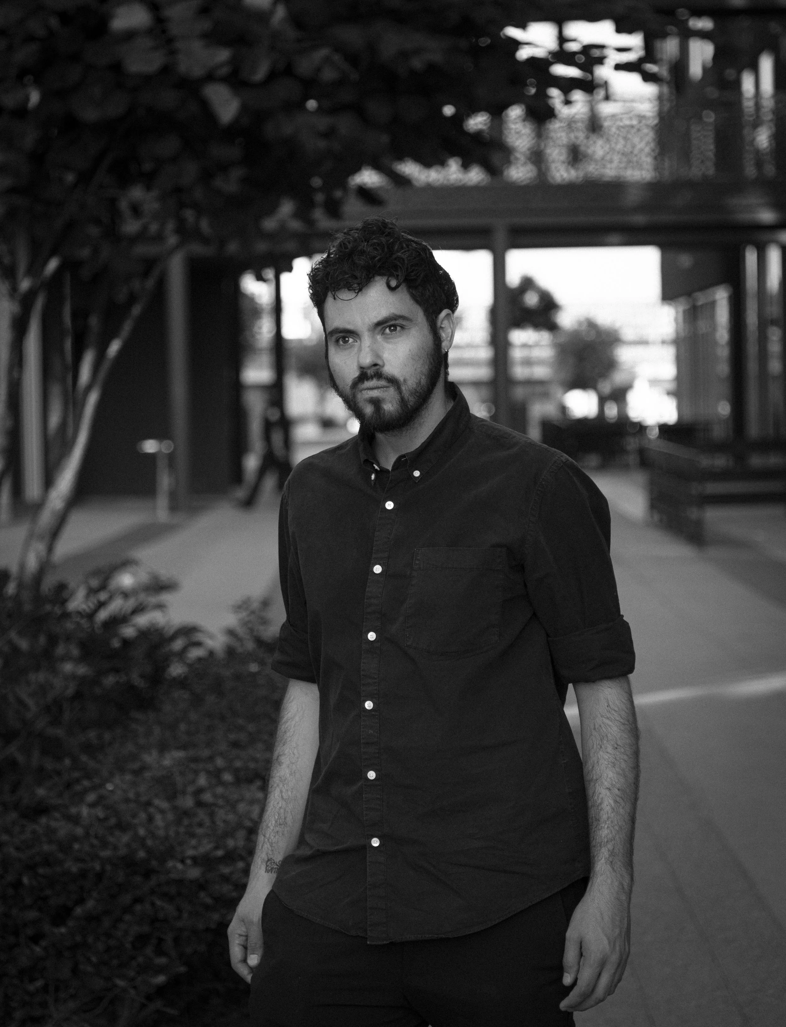 A young man with dark, curly hair and a beard, wearing a black button-up shirt with sleeves rolled up, standing outdoors in an urban setting with modern architecture and trees in the background.