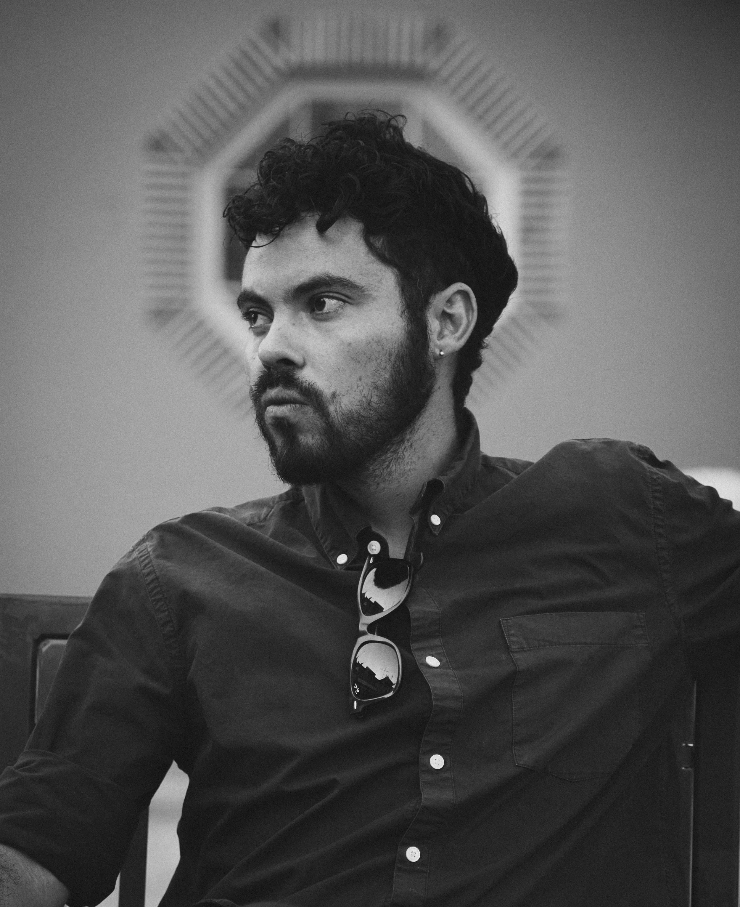 A man with dark, curly hair, a beard, and earrings sitting in front of a mirror with a decorative frame, wearing a button-up shirt and sunglasses hanging from the shirt collar.