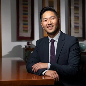 A man in a dark suit, white shirt, and purple tie sitting at a wooden table, smiling, in an indoor setting with framed artwork on the wall behind him.